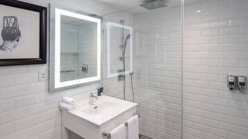 Ein modernes Badezimmer mit weiß gefliesten Wänden, einer Duschkabine aus Glas, einem weißen Waschbecken mit Spiegel und einem gerahmten Kunstwerk darüber.