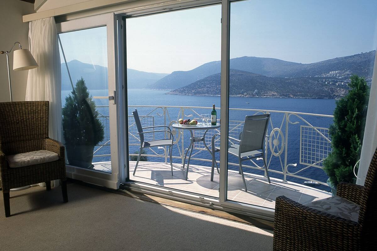 A room with a large sliding glass door leading to a balcony with a view of the sea and mountains. There are wicker chairs and a table with a bottle on it.