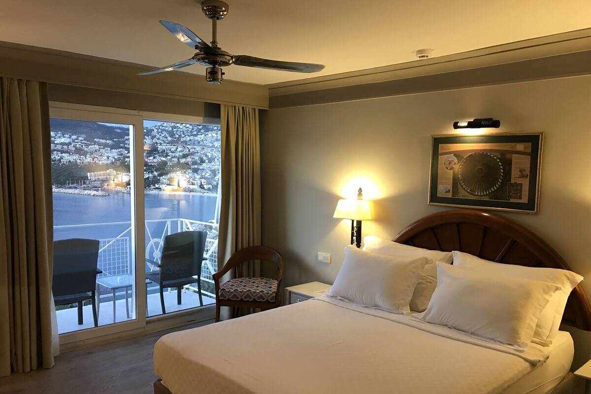 A hotel room with a bed, a chair, a nightstand, a lamp, a ceiling fan, and a large window with a view of a city and water.