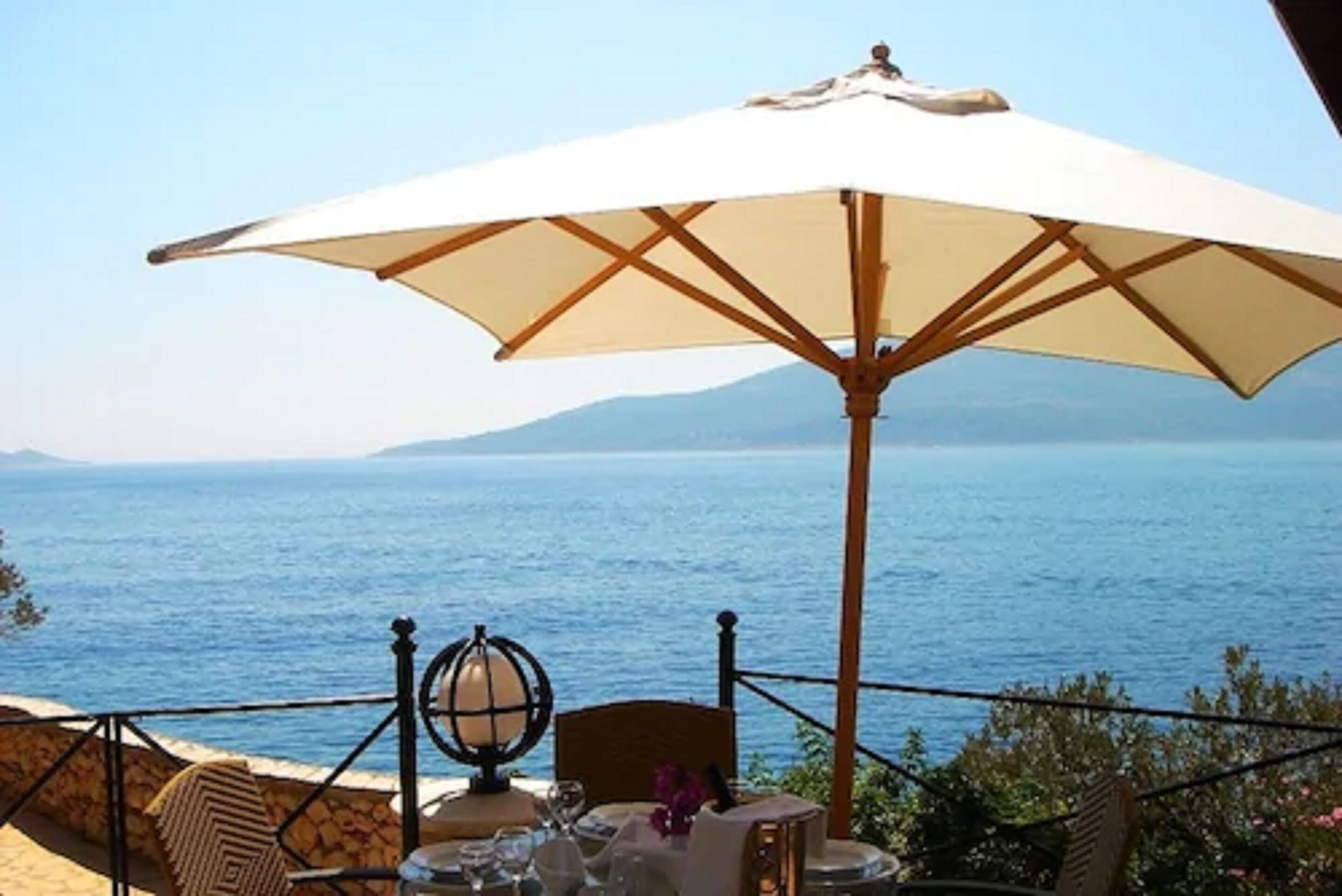 A table with a white umbrella, a globe, and a chair, overlooking a body of water with mountains in the background.
