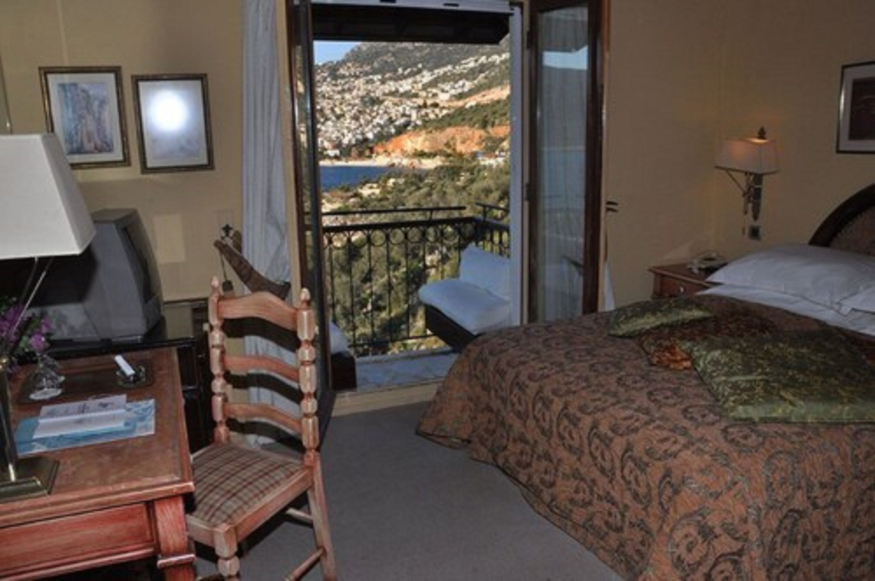 Ein Hotelzimmer mit Bett, Schreibtisch mit Lampe, Stuhl und Balkon mit Blick auf eine Hügel- und Wasserlandschaft.