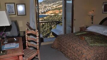 Ein Hotelzimmer mit Bett, Schreibtisch mit Lampe, Stuhl und Balkon mit Blick auf eine Hügel- und Wasserlandschaft.