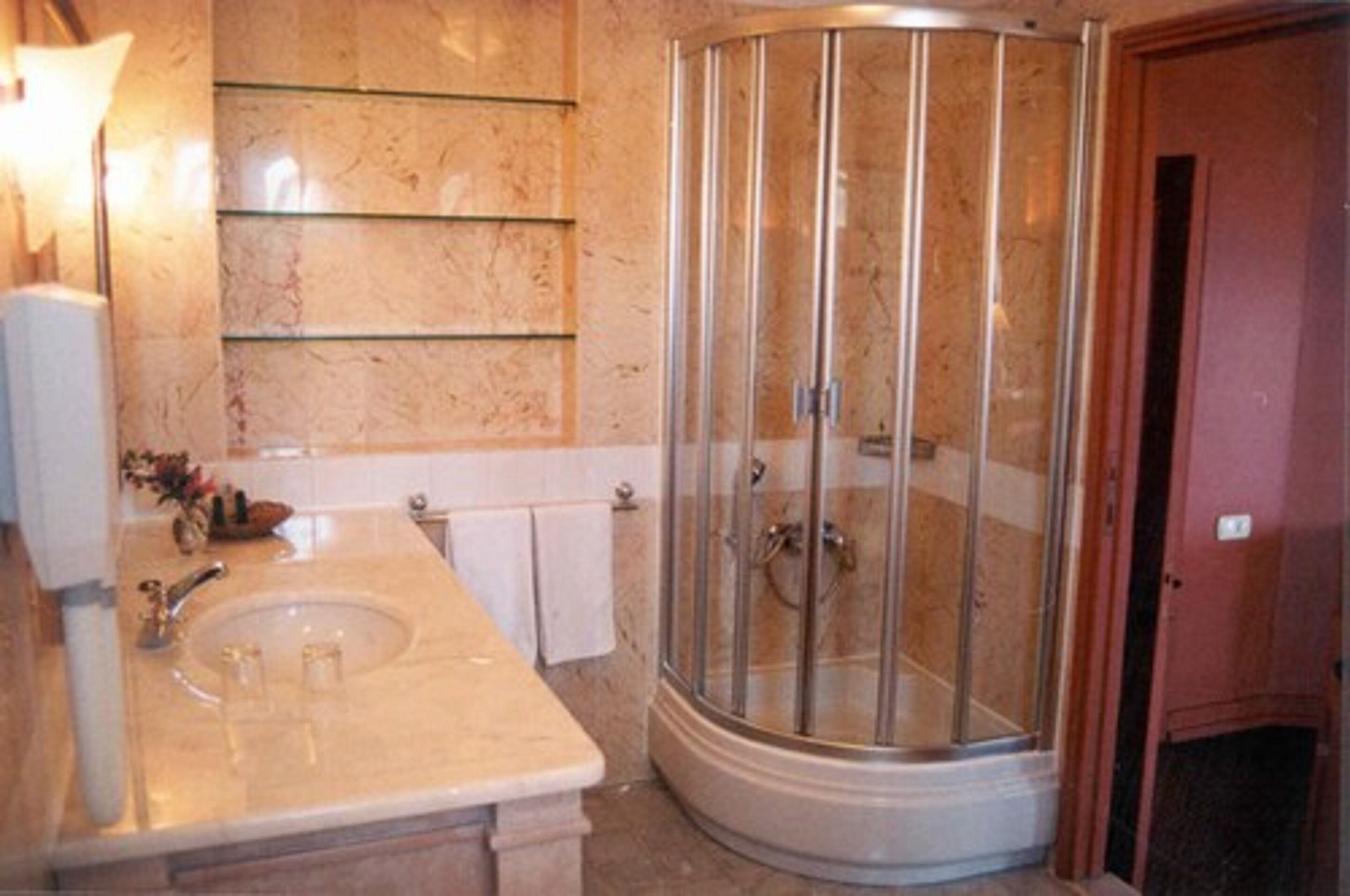 A bathroom with a marble sink, glass shelves, a glass-enclosed shower, and a white door.