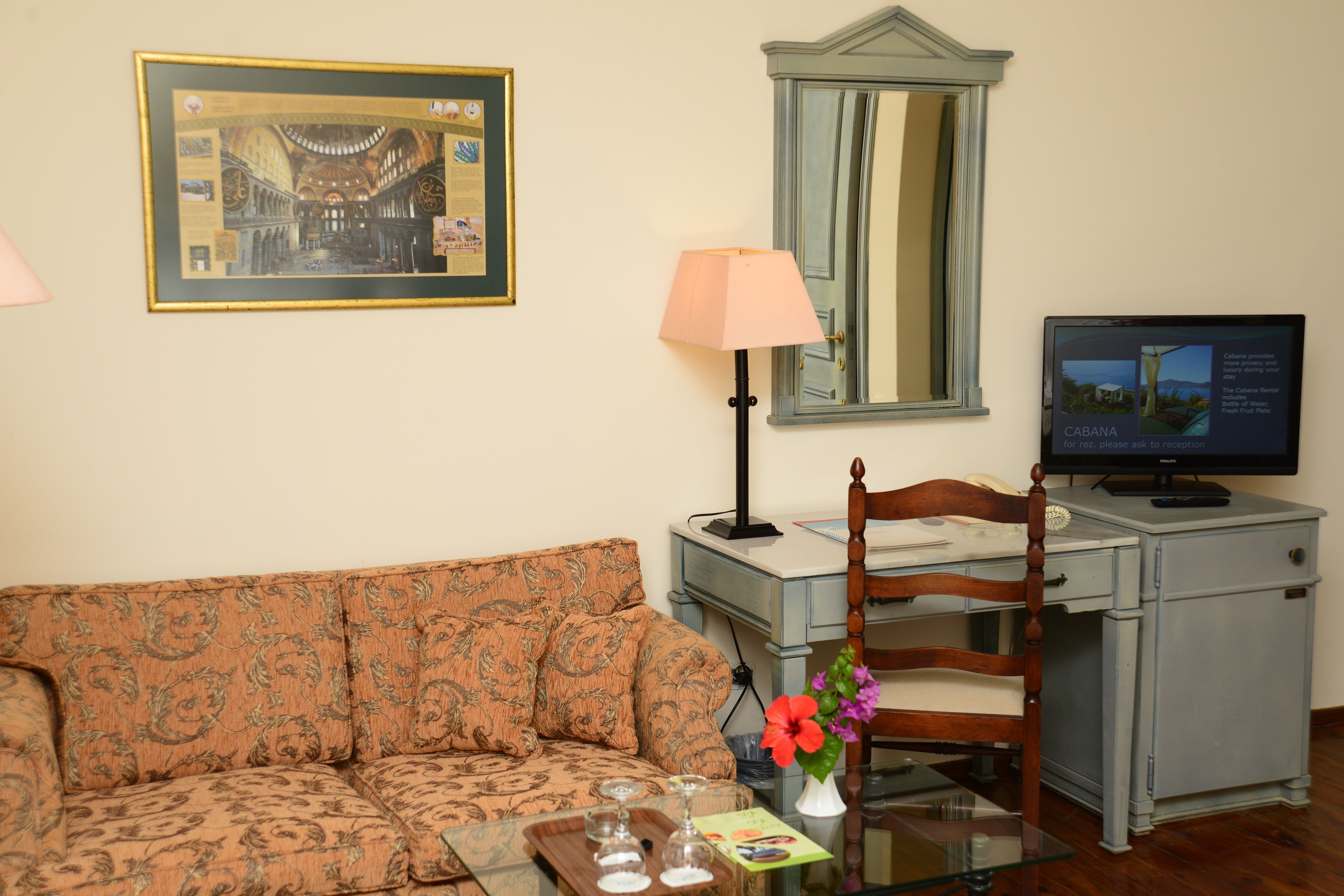 Una sala de estar con un sofá, una mesa con vasos, una lámpara, una silla, un escritorio con una computadora y un cuadro en la pared.