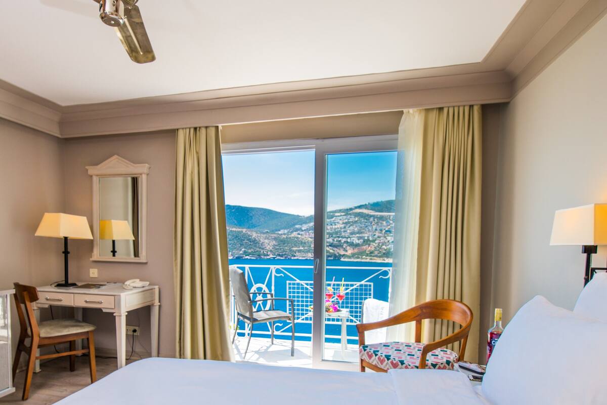 A hotel room with a bed, desk, chair, and a balcony with a view of the landscape.