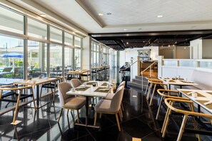 Un interior de restaurante moderno con grandes ventanales, pisos de baldosas negras y una combinación de mobiliario de madera y metal.