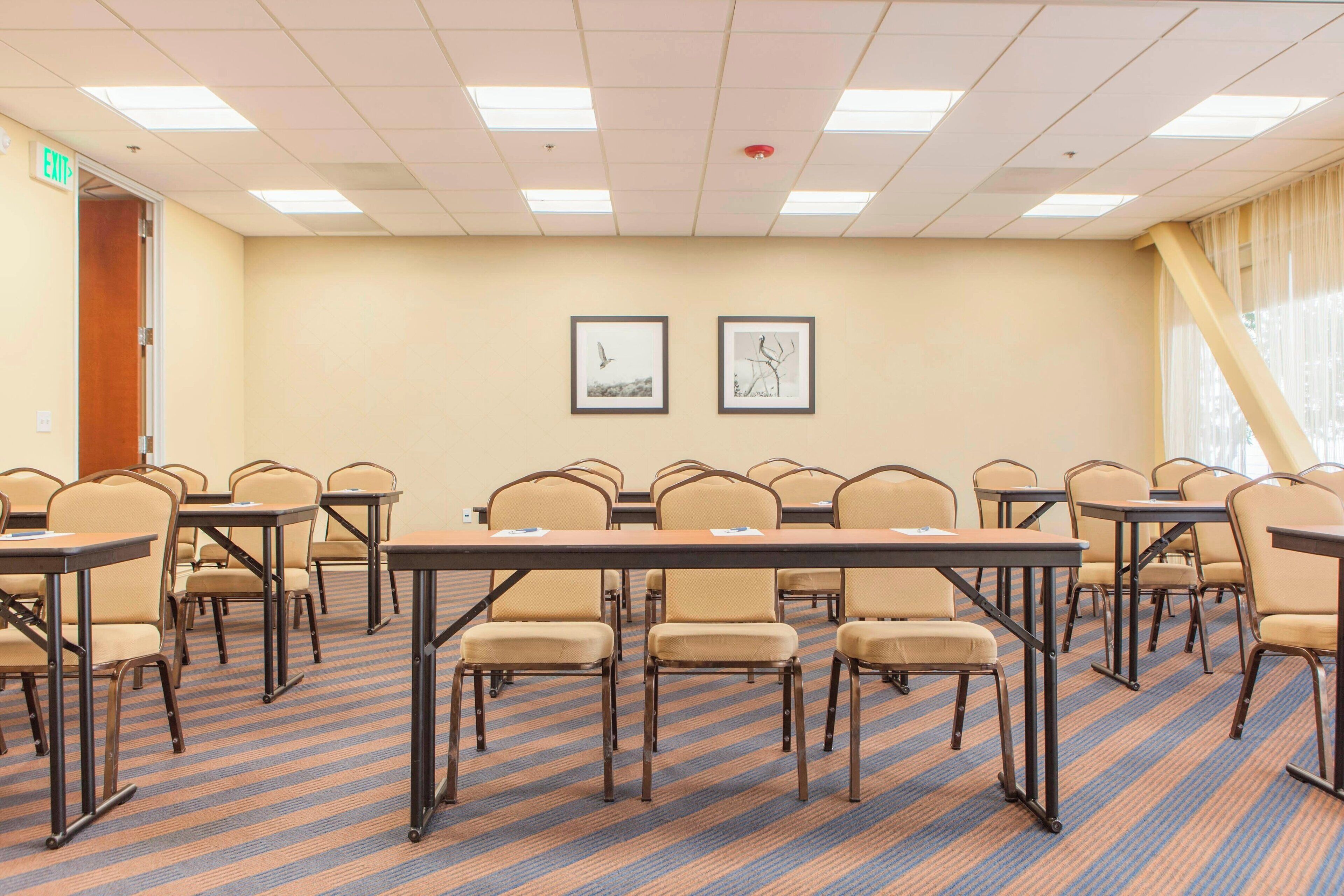 A conference room with tables and chairs arranged for meetings. There are framed pictures on the wall, a door labeled "EXIT", and a window with curtains.