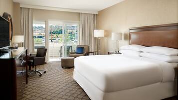 A hotel room with a large bed, a desk with a TV, a chair, and a view of a scenic landscape through the window.
