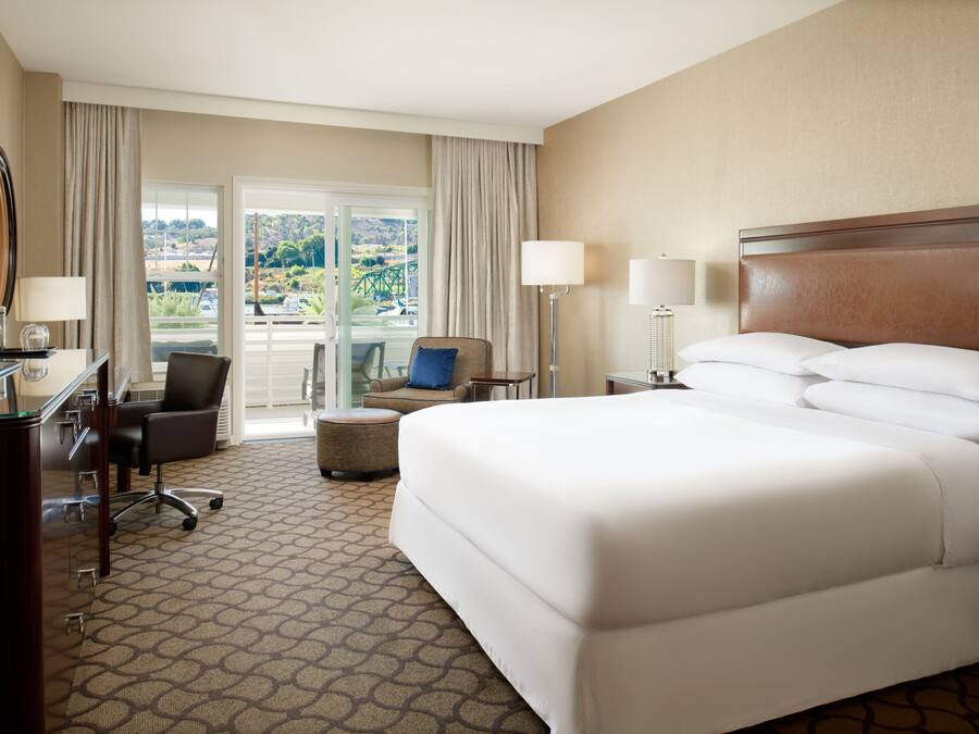 A hotel room with a large bed, a desk with a TV, a chair, and a view of a scenic landscape through the window.