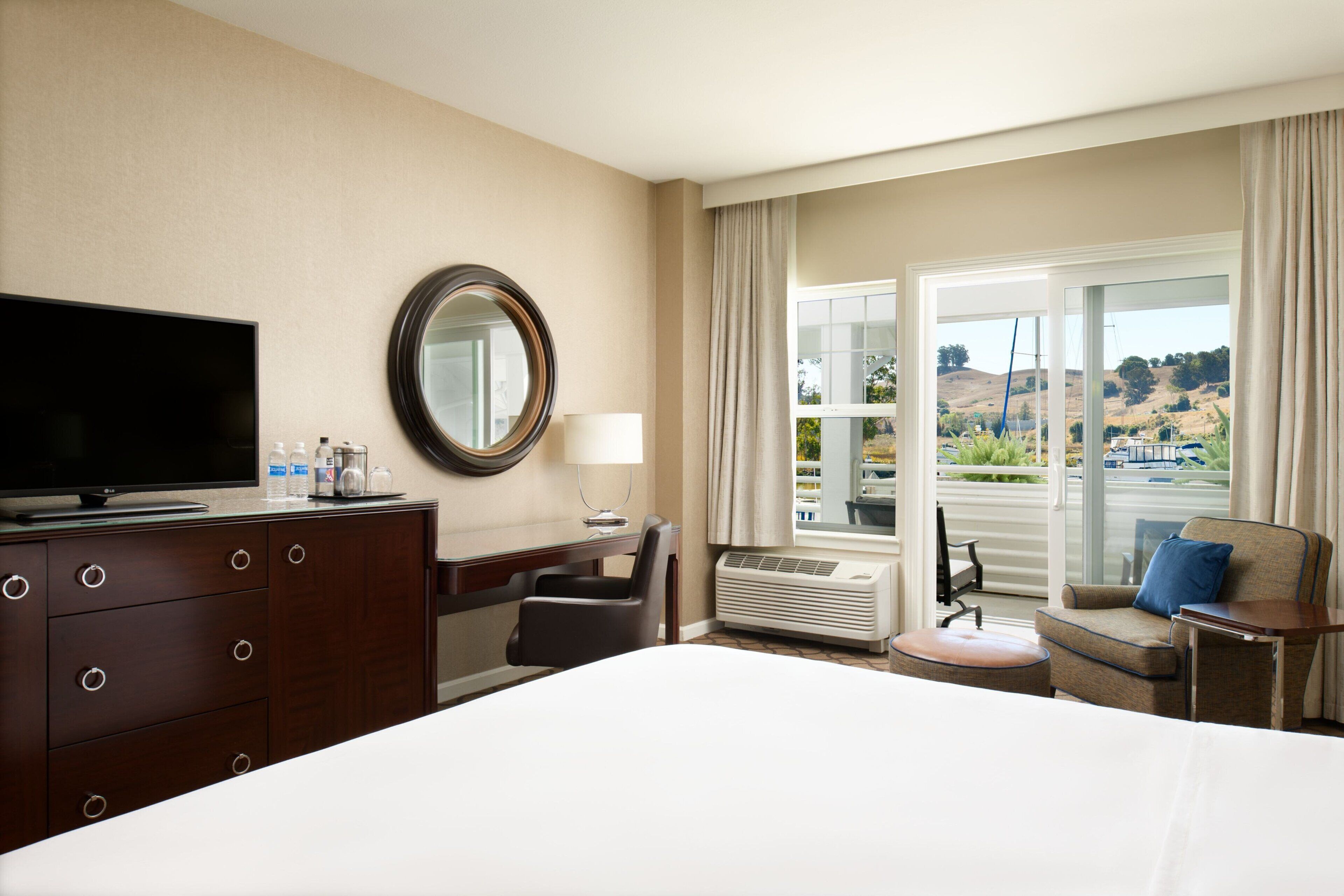 Ein Hotelzimmer mit Bett, Fernseher, Schreibtisch mit Stuhl, Fenster mit Blick auf Hügel und einem großen Spiegel.