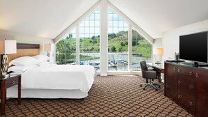 Habitación de hotel con una cama grande, un escritorio con televisor, una silla y vistas a un puerto deportivo a través de amplios ventanales.