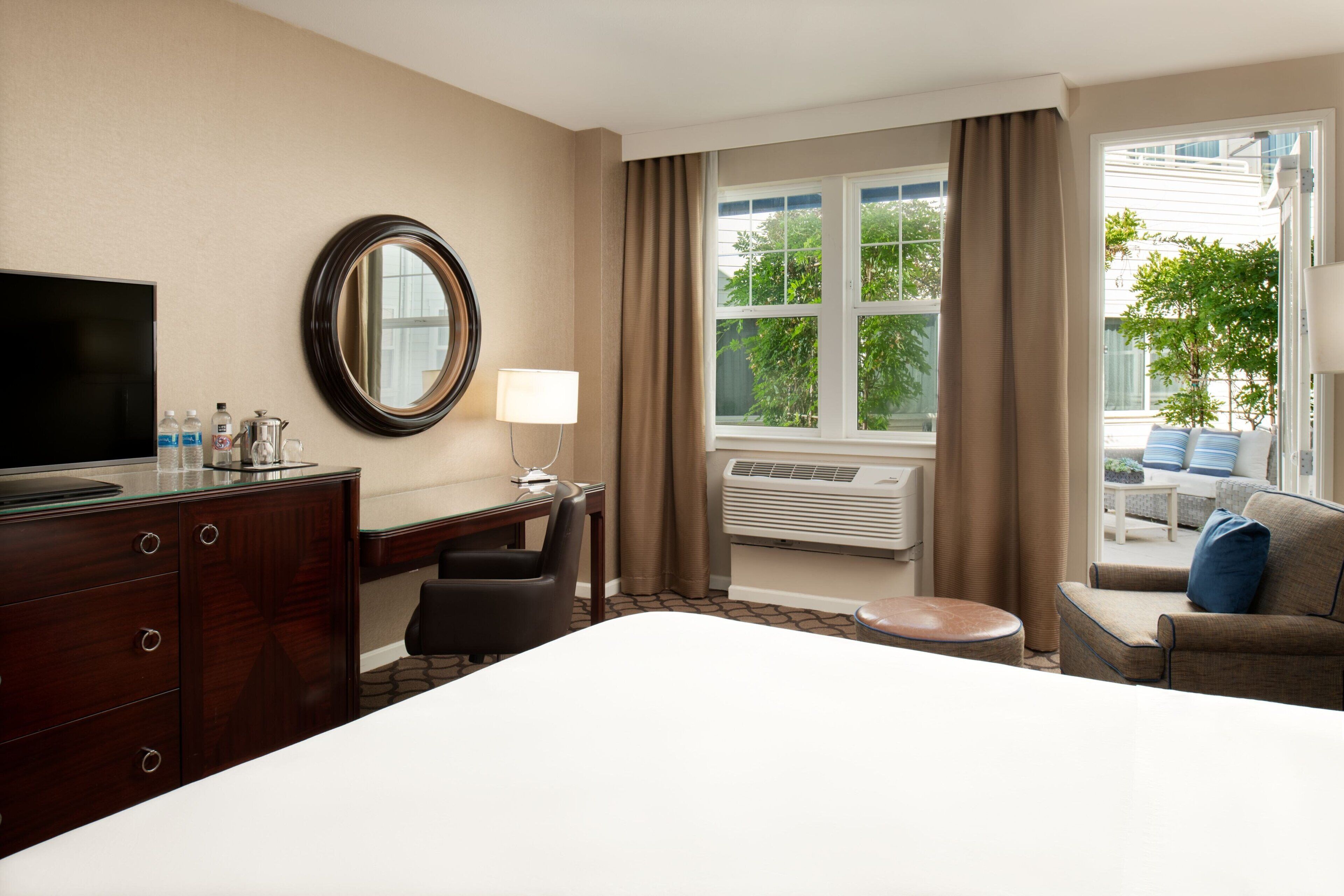 Ein Hotelzimmer mit Bett, Schreibtisch mit Stuhl, Fernseher, Spiegel, Lampe, Klimaanlage und Blick ins Freie.