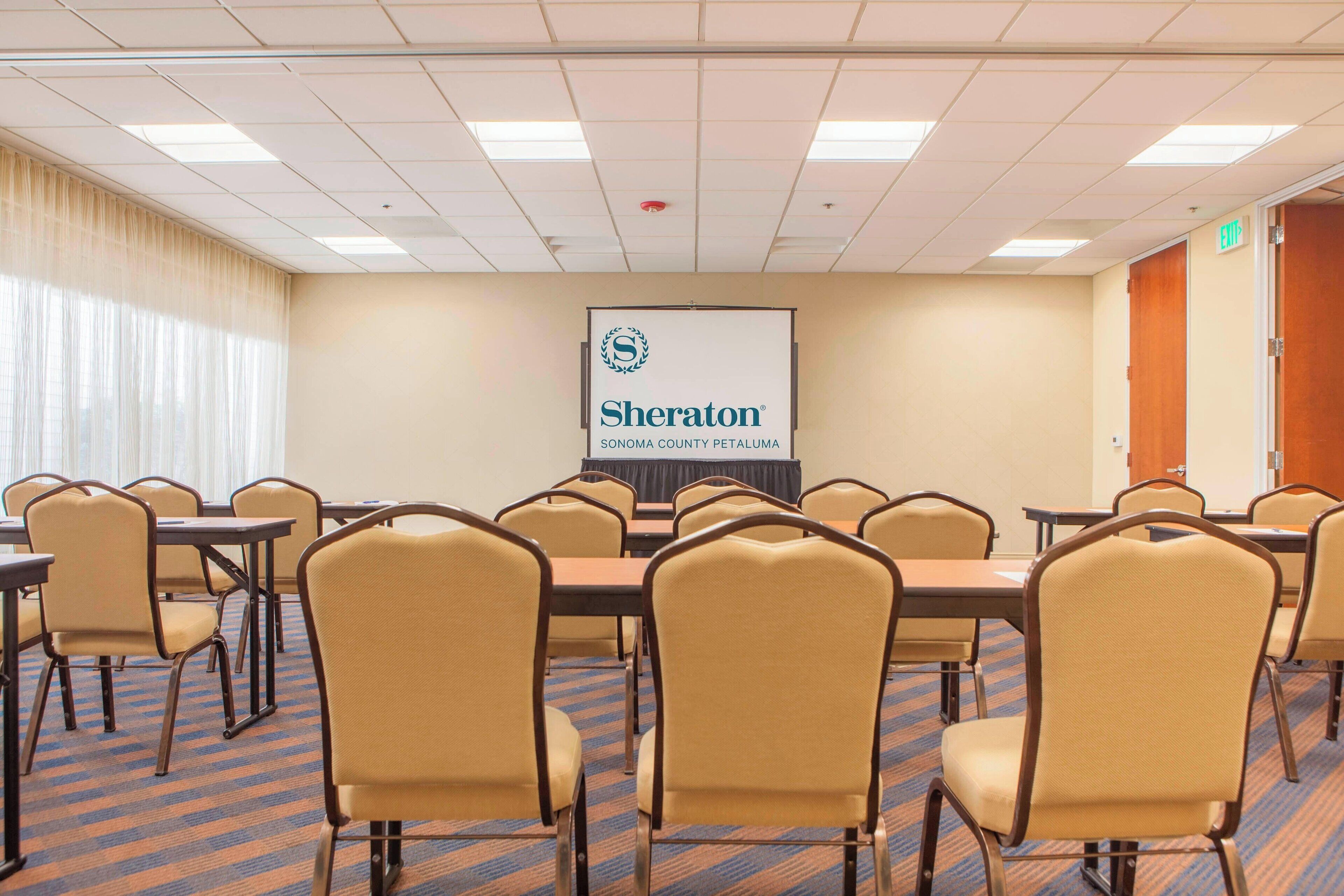Ein Konferenzraum mit beigefarbenen Stühlen und Tischen, einer Projektionsleinwand mit dem Sheraton-Logo und einem Teppichboden.