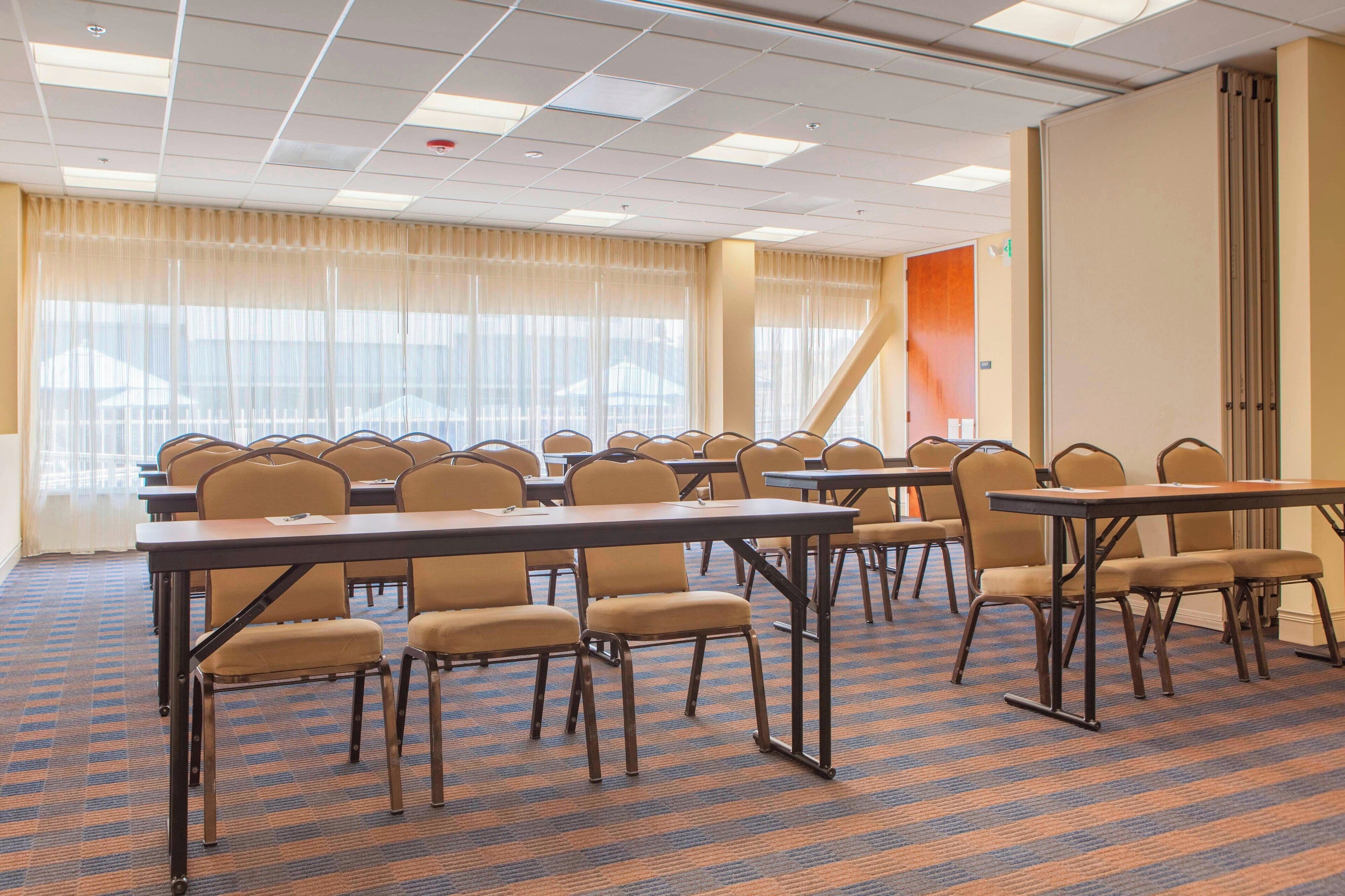 A conference room with multiple tables and chairs, large windows with sheer curtains, and a carpeted floor.