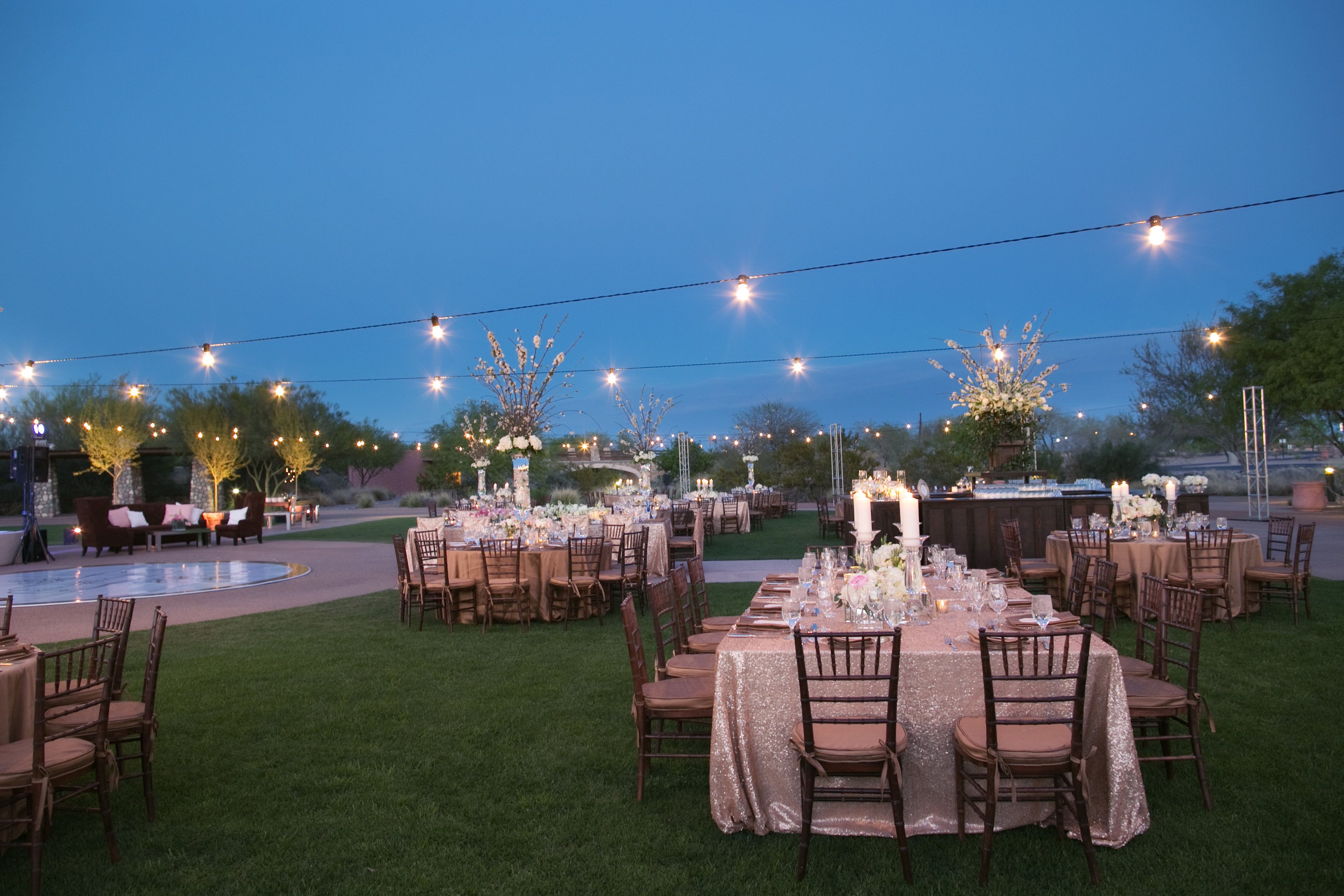 An outdoor event space with round tables and chairs, string lights, and floral arrangements.