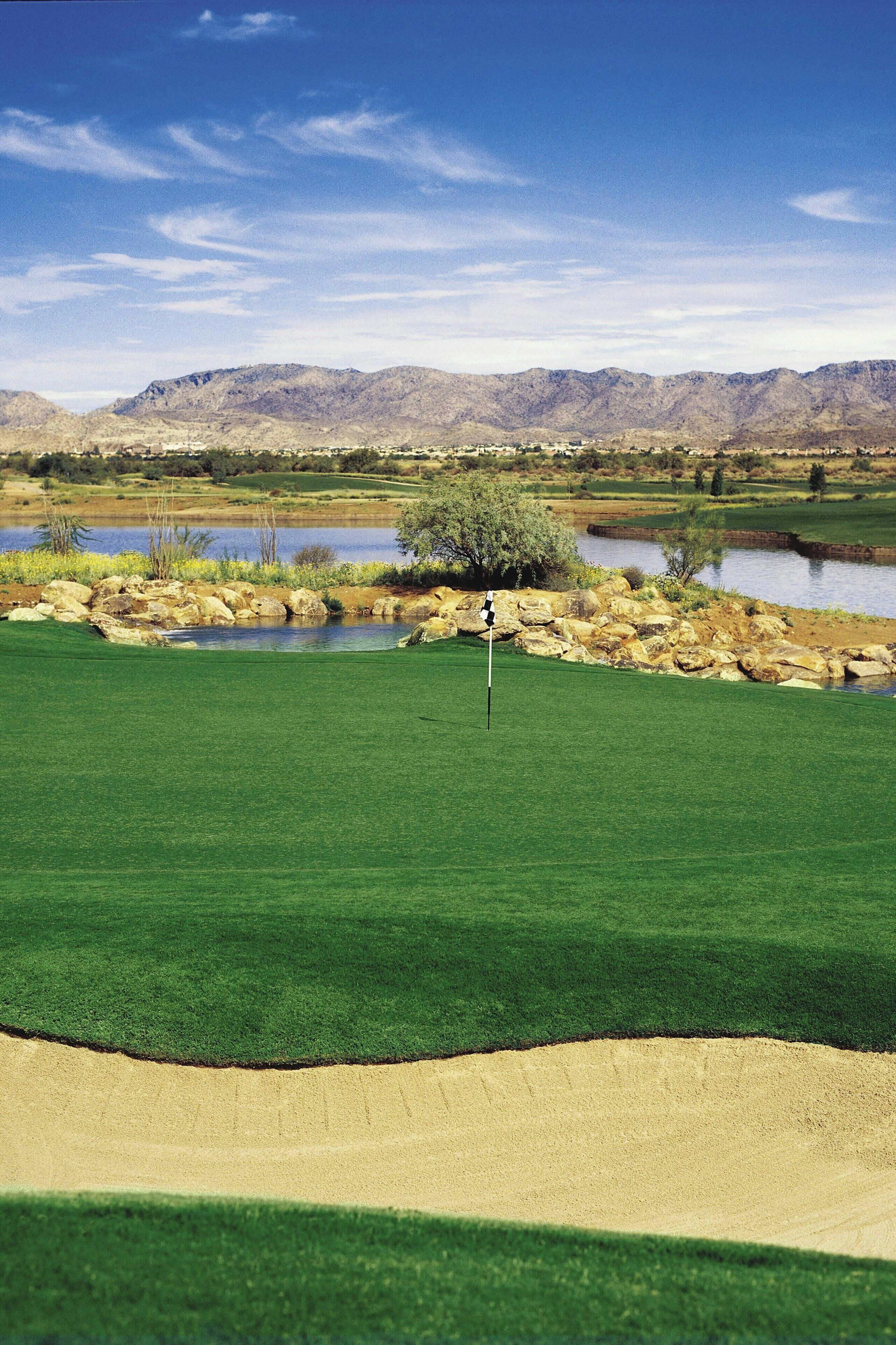 Un campo de golf con un marcador de bandera, peligros de agua y montañas a lo lejos.