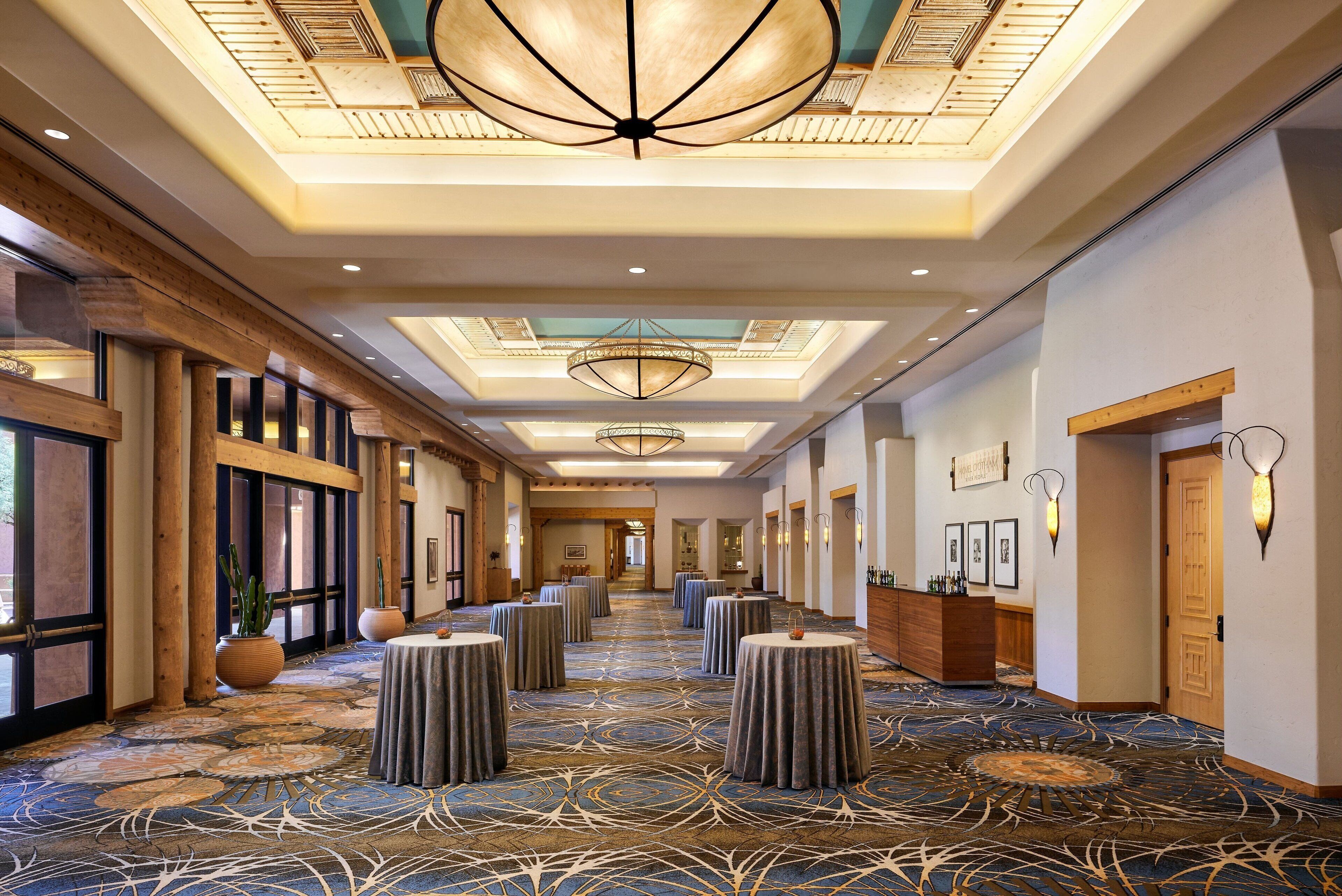 A hotel corridor with round tables, a reception desk, and a high ceiling with chandeliers.