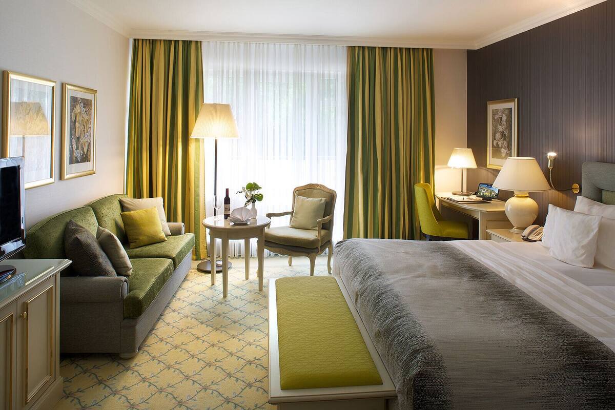 A hotel room with a bed, a sofa, a chair, a small table, a TV, and a lamp.