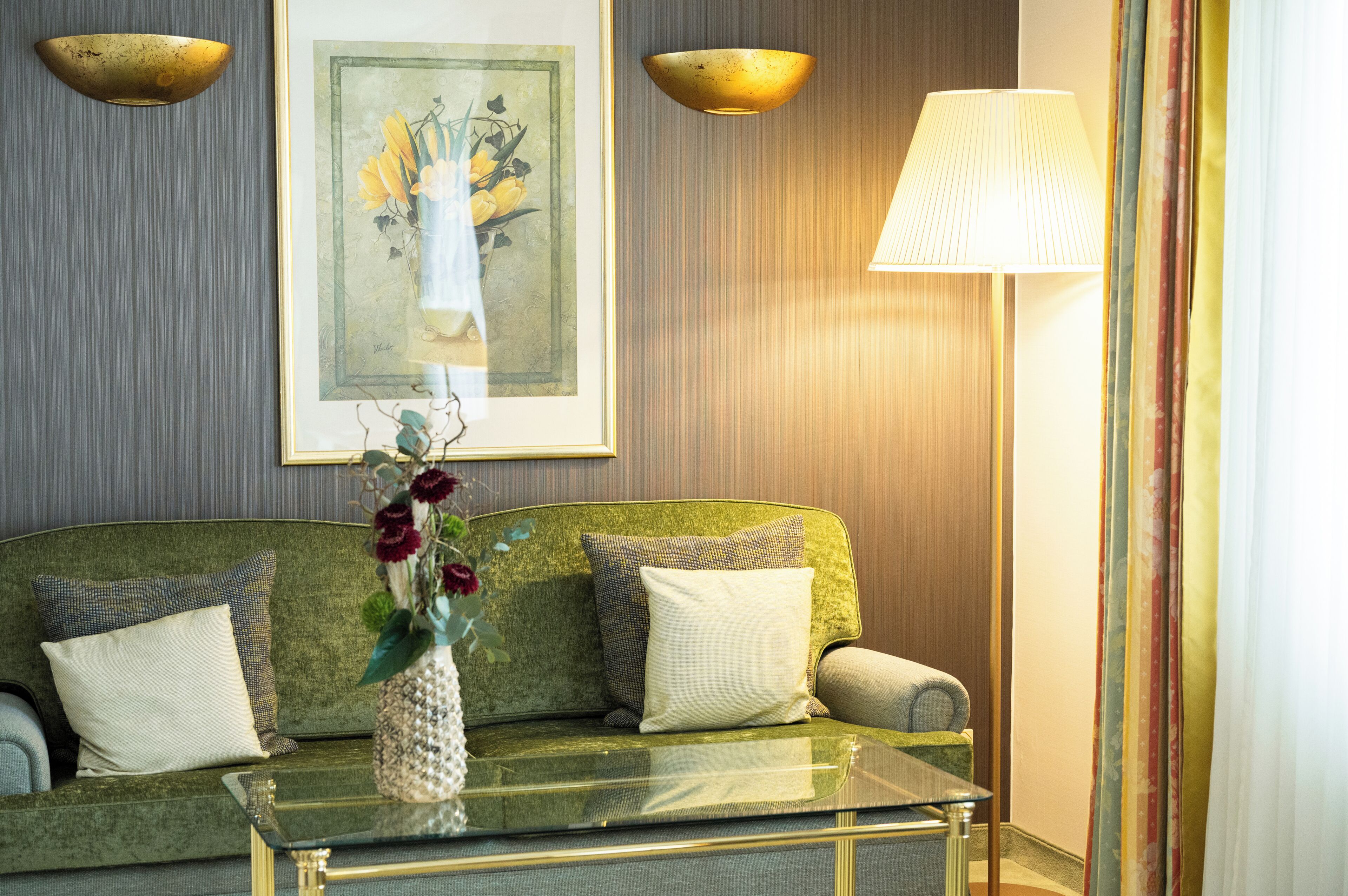 A green sofa with two pillows, a glass coffee table, a vase with flowers, a framed artwork on the wall, and a lamp.