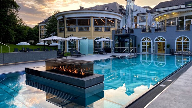 A pool area with a fire feature, lounge chairs, and umbrellas. - Der Sonnenhof (Bad Woerishofen)