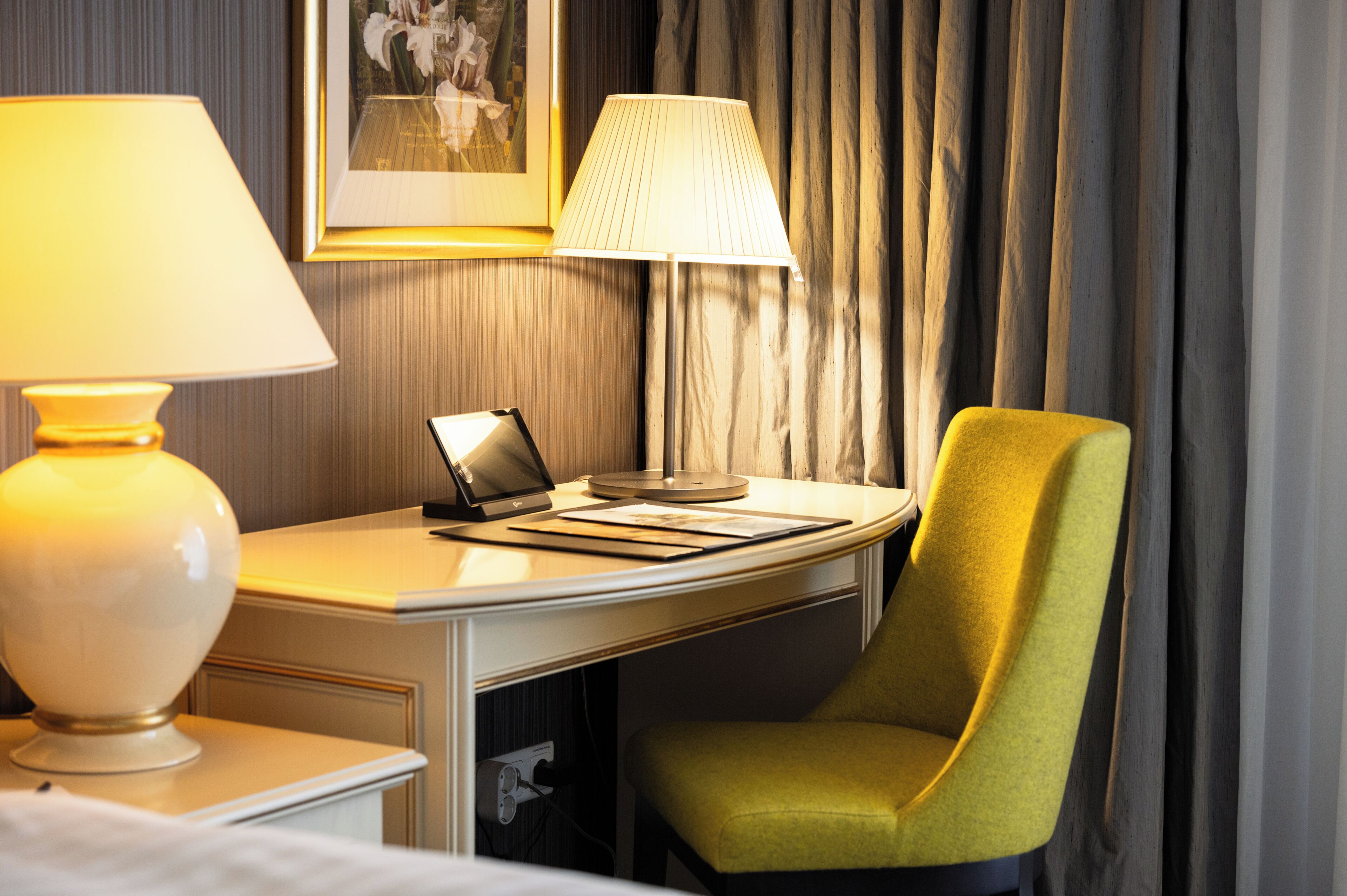 A hotel room with a desk, two lamps, a laptop, a chair, and a framed picture on the wall.