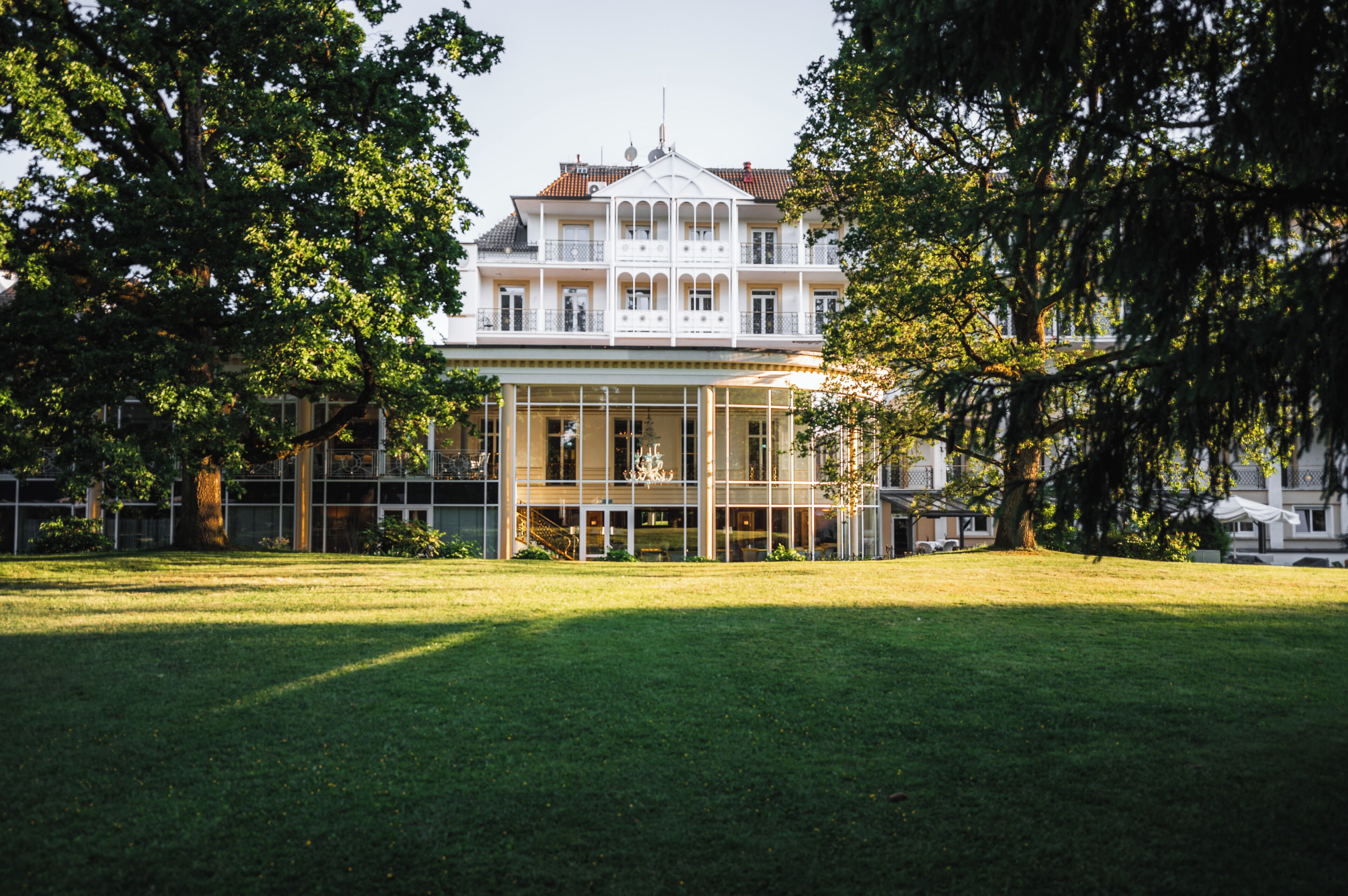 A large, multi-story building with a central glass facade, surrounded by a well-maintained lawn and mature trees.