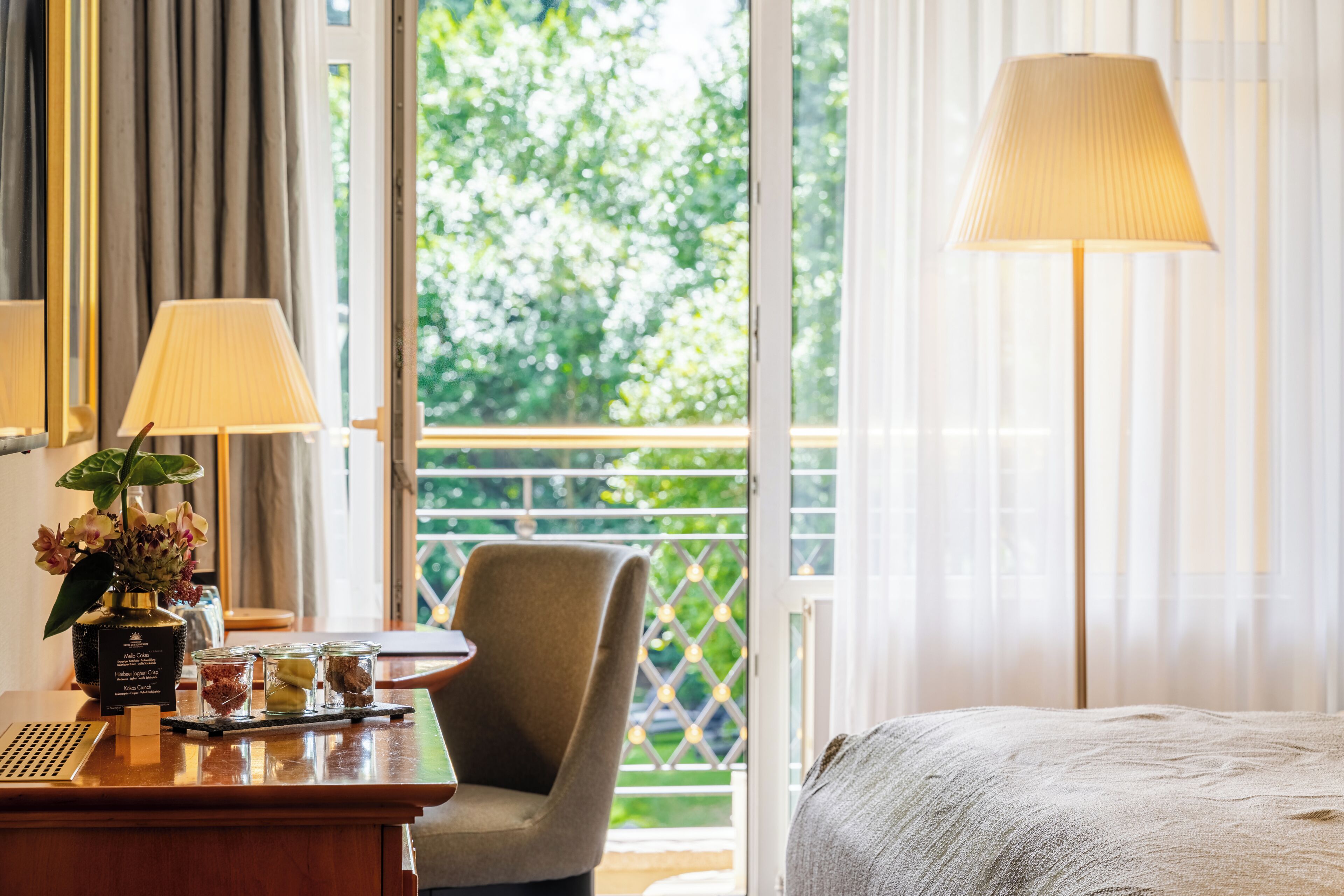 A hotel room with a bed, a desk with a lamp, a chair, and a view of greenery outside.
