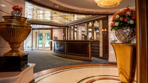 A lobby with a reception desk, a large gold vase with flowers, a chandelier, and a glass ceiling. - Der Sonnenhof (Bad Woerishofen)