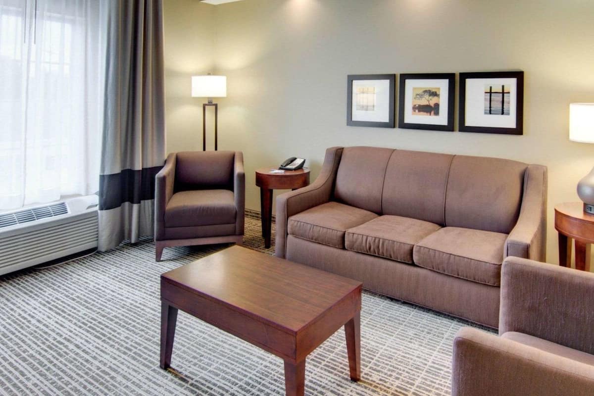 A hotel room with a sofa, armchair, and a small table. There are three framed pictures on the wall and a lamp on a side table.