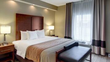 Een hotelkamer met een groot bed, twee bedlampen, een bankje en een raam met gordijnen.