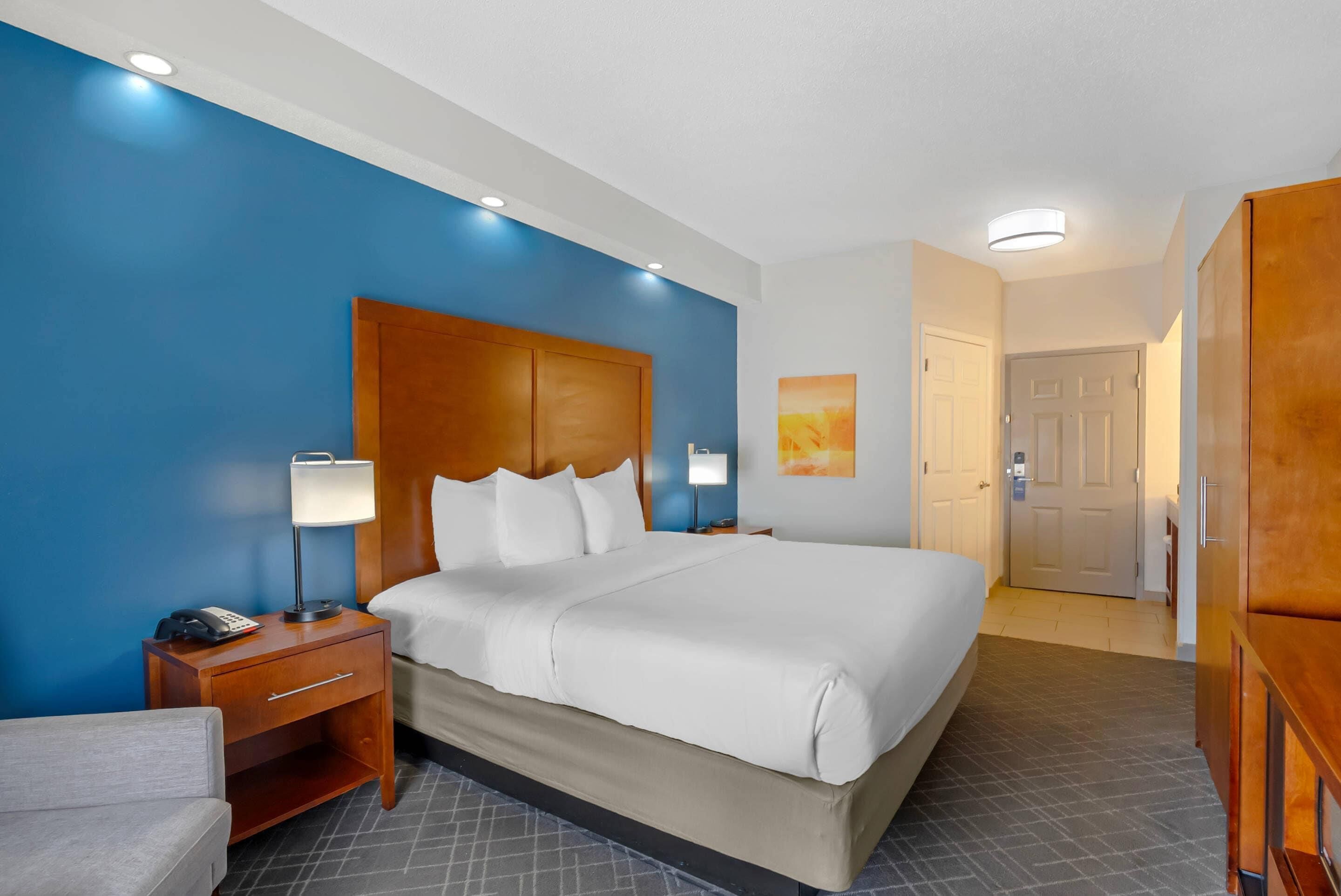 A hotel room with a large bed, a wooden nightstand with a lamp, a desk with a phone, and a door leading to another room.