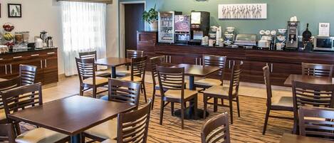 A hotel dining area with multiple tables and chairs, a counter with various food and beverage items, and a wall with a framed artwork.