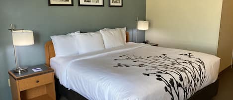 Una cama bien hecha con ropa blanca y un estampado floral negro, una mesita de noche de madera con una lámpara, y tres cuadros en la pared.