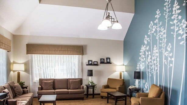 Una sala de estar con sofá, sillones, una mesa de centro y una alfombra estampada. Hay dos lámparas, una ventana con cortinas y una pared con diseño floral.