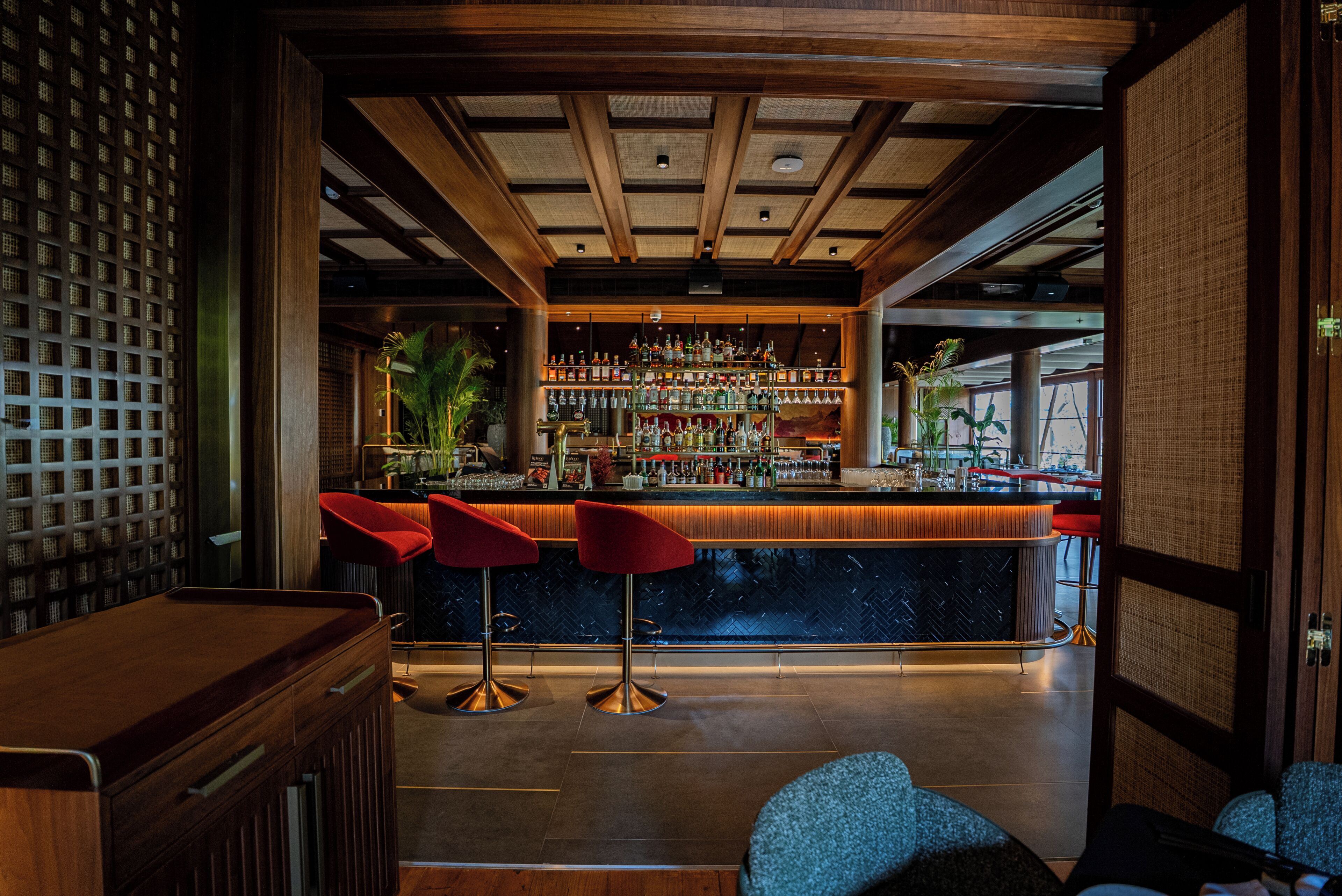 A bar area with a wooden ceiling, red bar stools, and a variety of bottles and glasses on shelves.