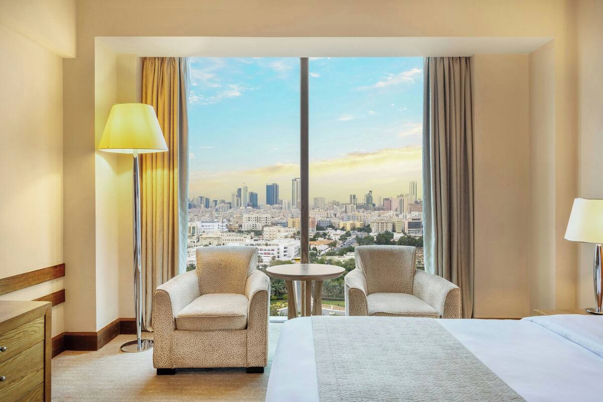 A hotel room with a large window, two armchairs, a bed, a round table, and a lamp.