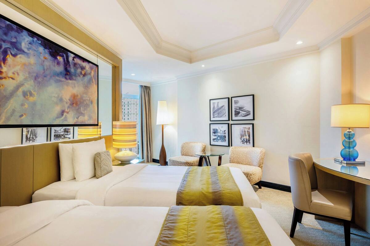 A hotel room with two beds, a desk, a chair, a lamp, and framed pictures on the wall.