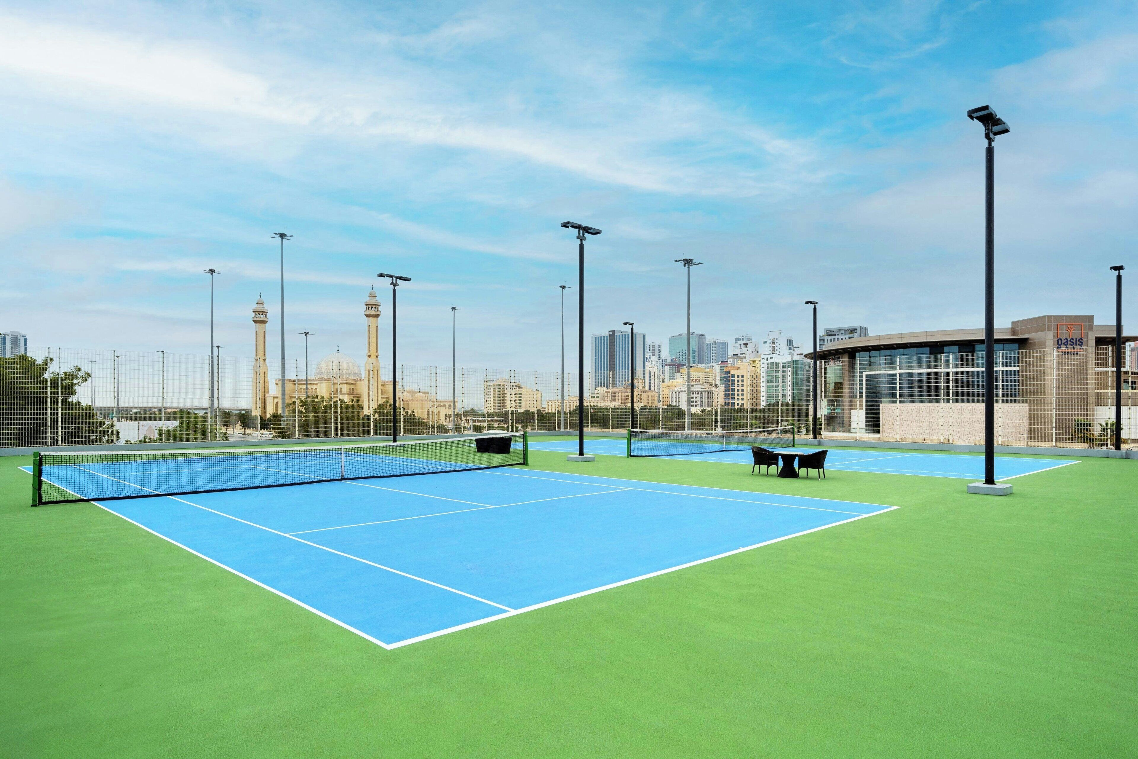 Un court de tennis en plein air, doté d’un filet et entouré de gazon verdoyant, avec une vue sur la ville en arrière-plan.