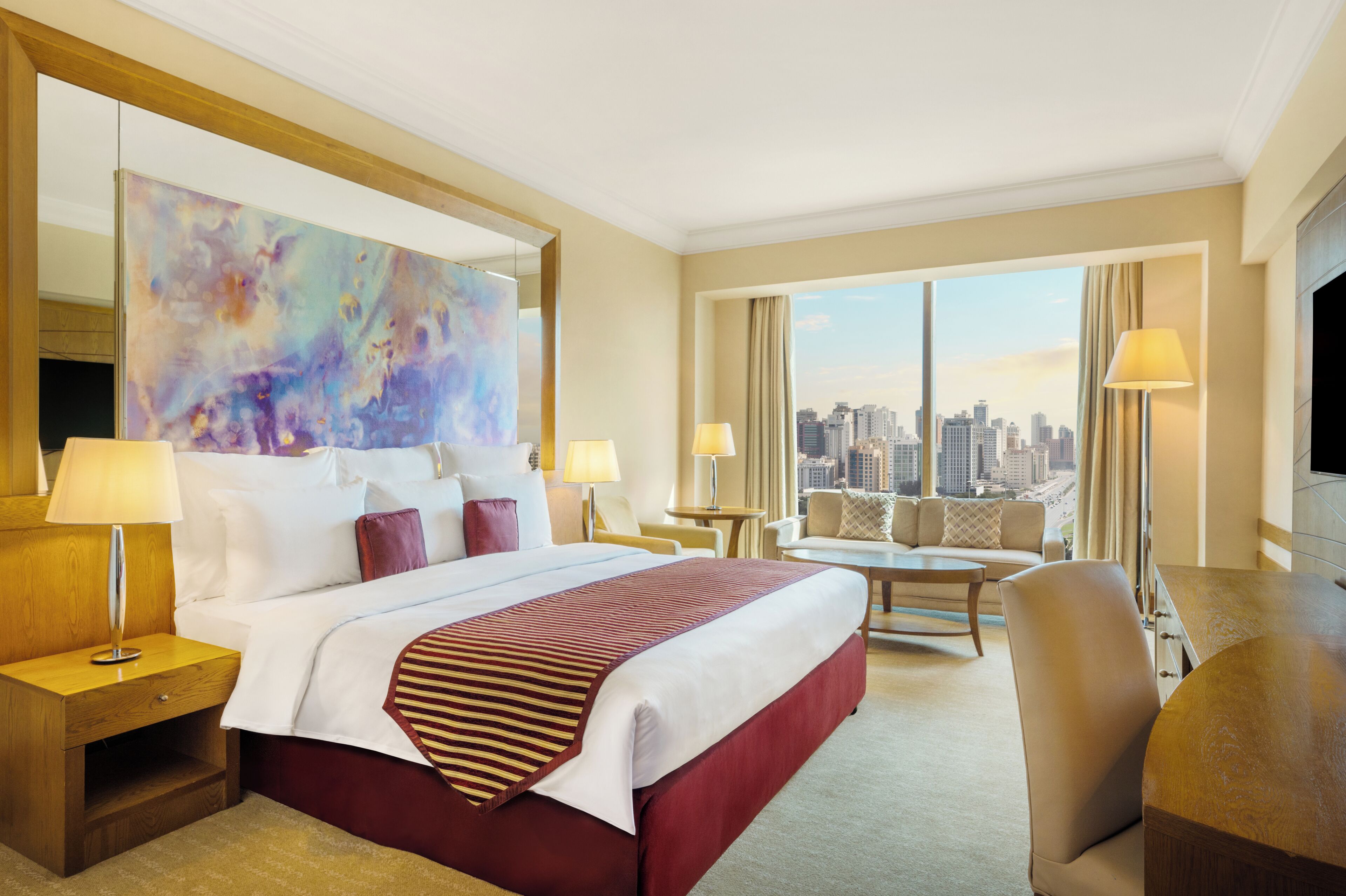 A hotel room with a large bed, a desk, a chair, and a television. There is a large window with a city view.