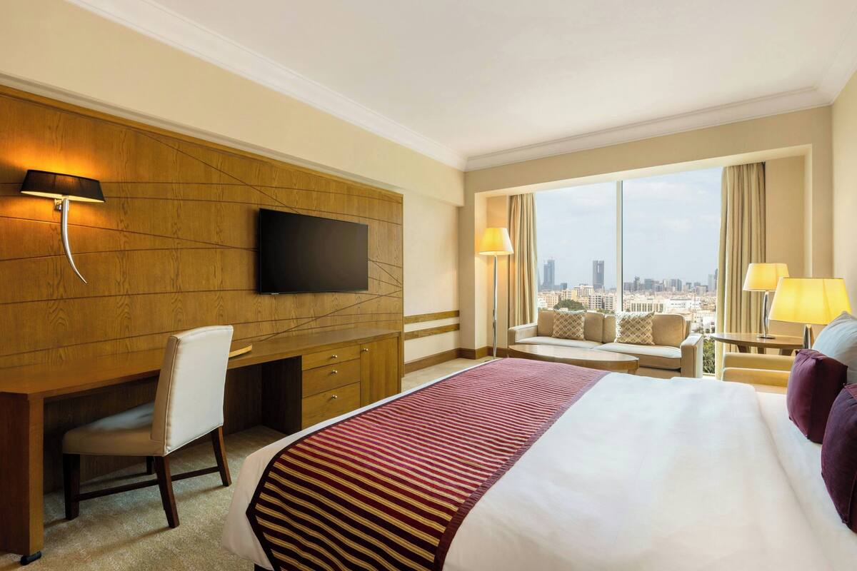 A hotel room with a large bed, a desk, a TV, and a view of the city.