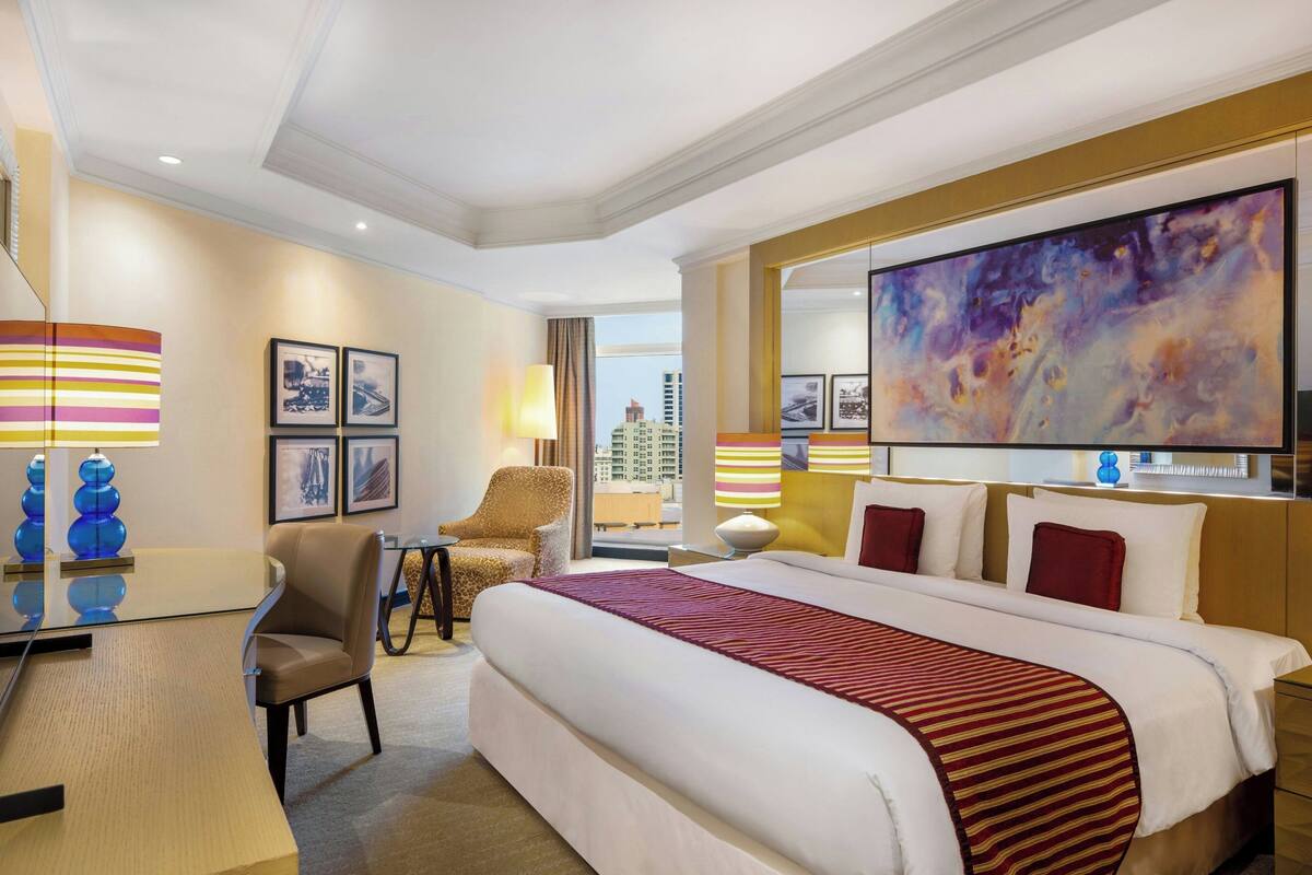 A hotel room with a large bed, a desk, a chair, and a large window offering a city view.