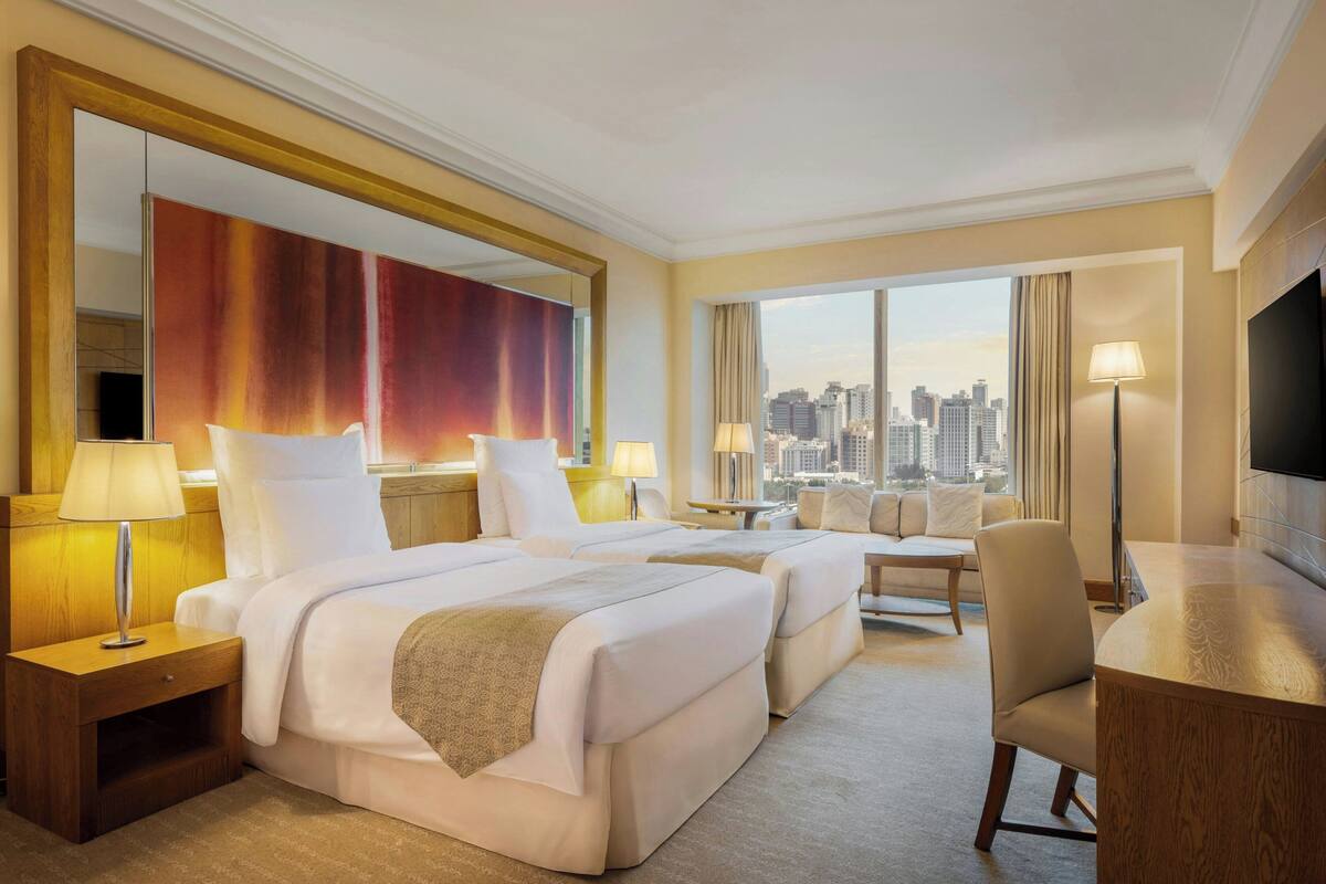 A hotel room with two beds, a desk, a chair, and a large window offering a city view.