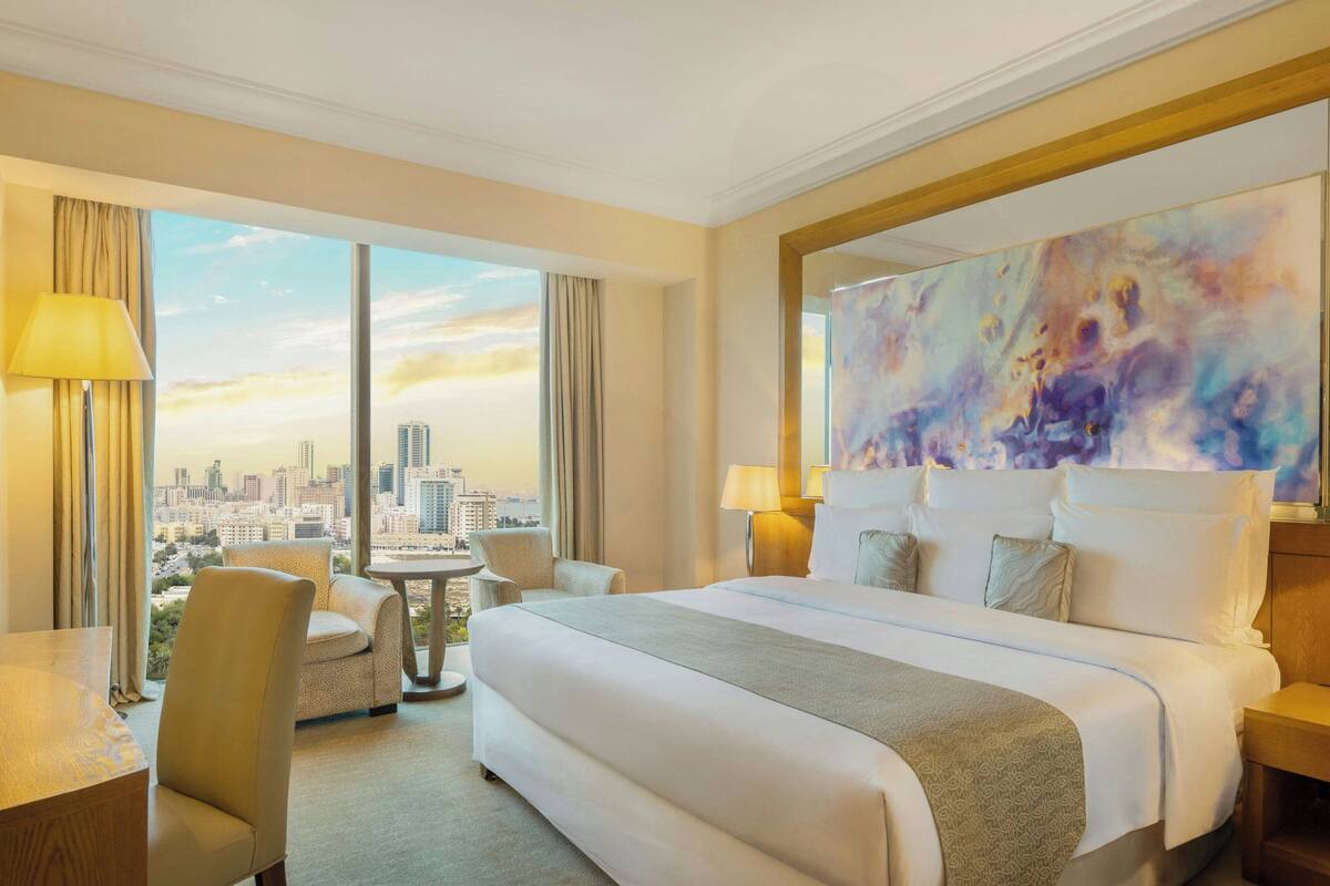 A hotel room with a large bed, a desk, a chair, and a view of the city skyline through large windows.