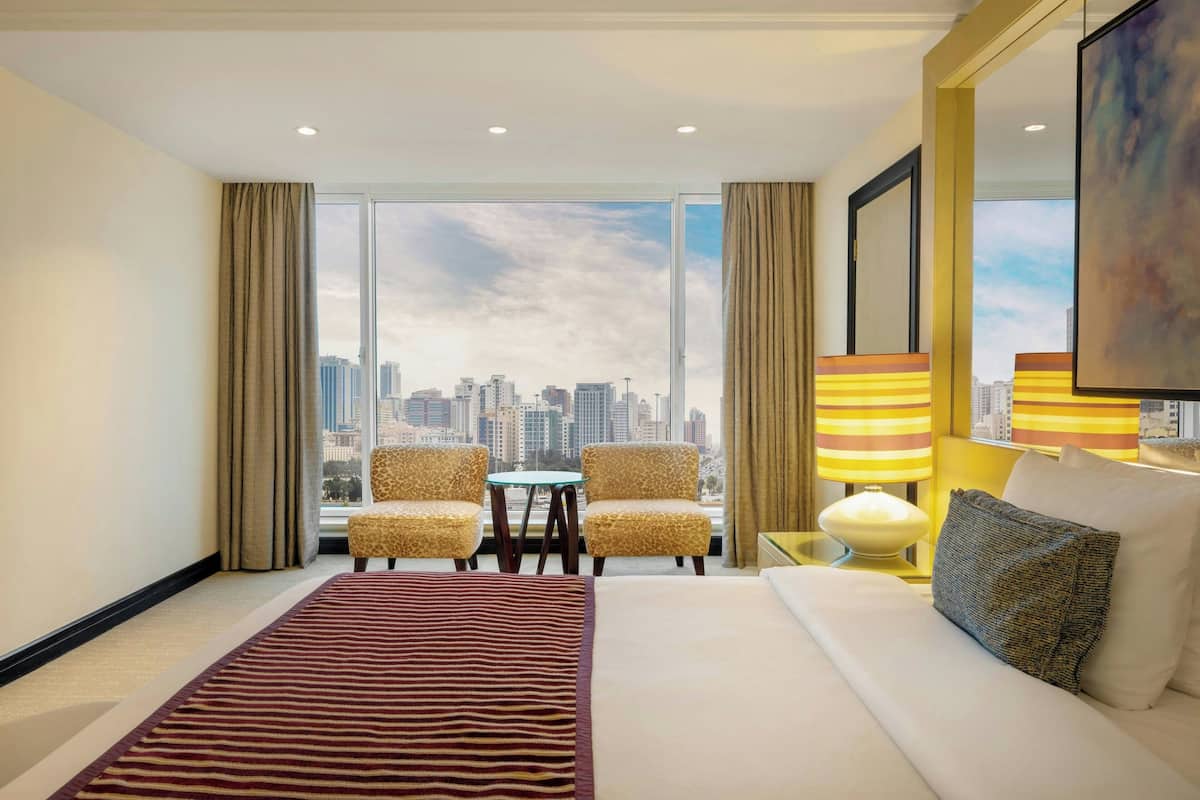 A hotel room with a large window, a bed, a sofa, two armchairs, a small table, and a lamp.