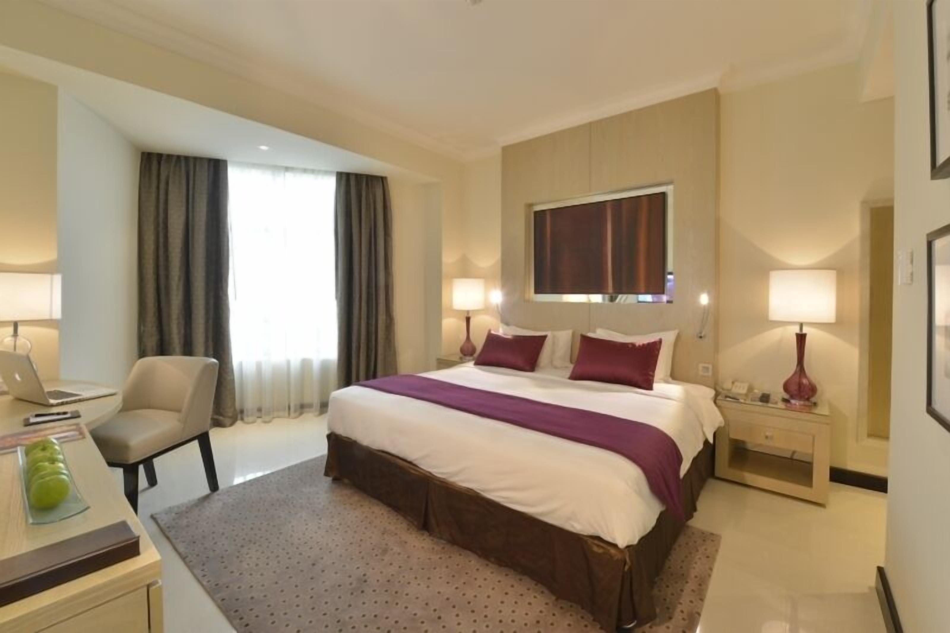 A hotel room with a bed, two bedside tables, a desk with a chair, a window with curtains, and a lamp.