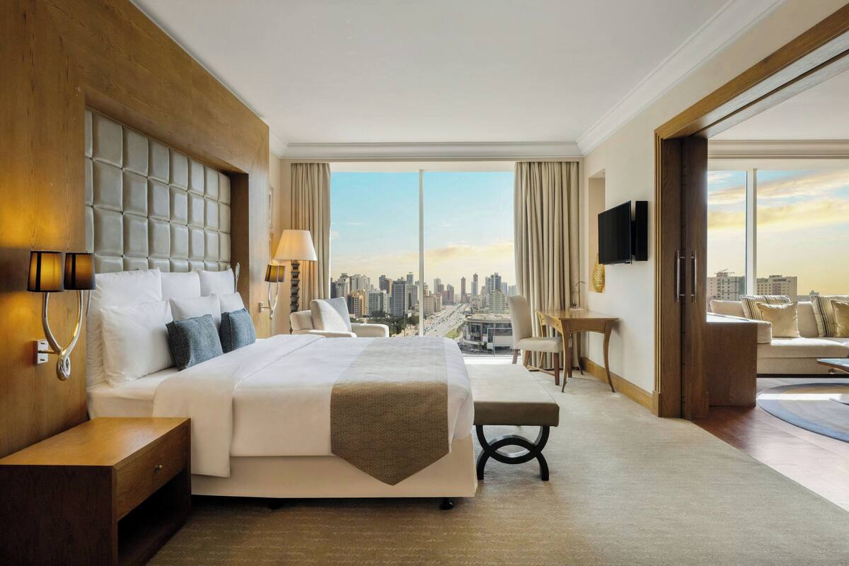 A modern hotel room with a large bed, a desk, a chair, and a view of the city skyline.