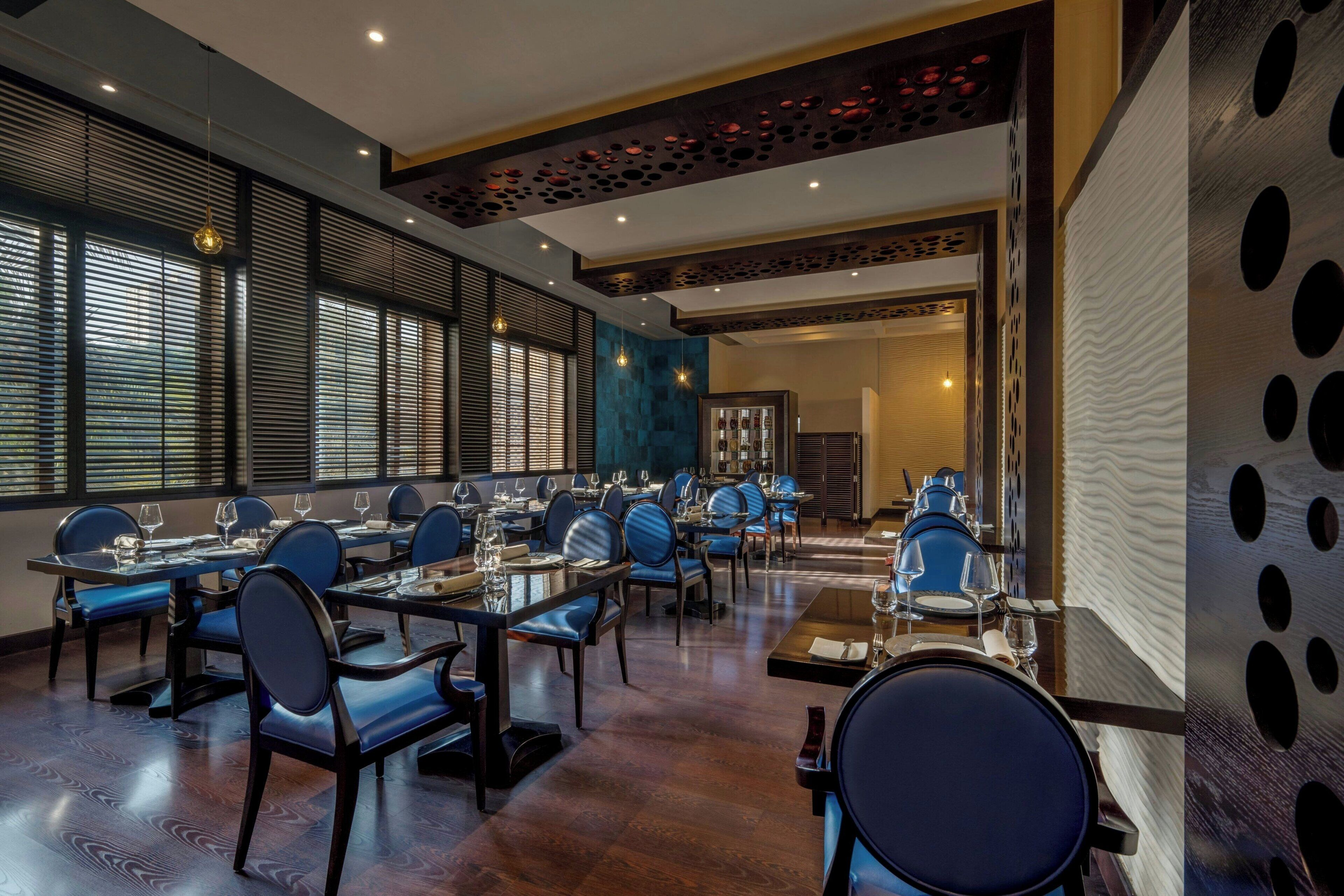 A dining area with multiple tables and chairs, large windows with blinds, and a decorative ceiling.