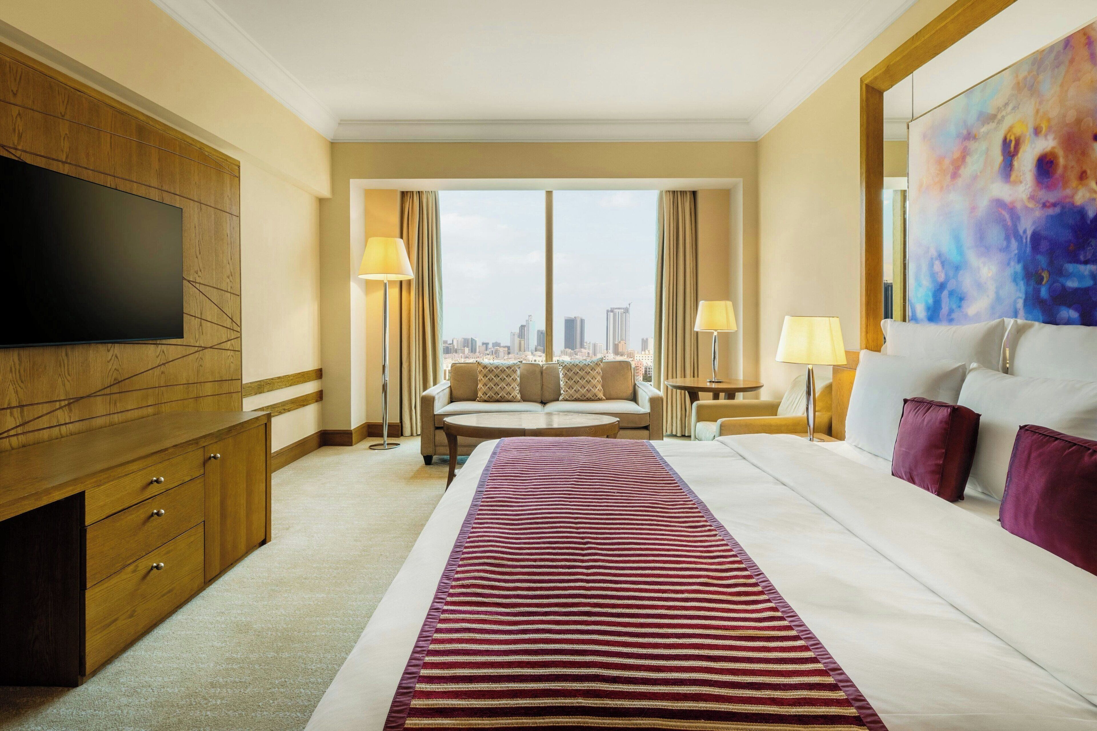 A hotel room with a large bed, a flat-screen TV mounted on the wall, a sofa, two lamps, and a city view.