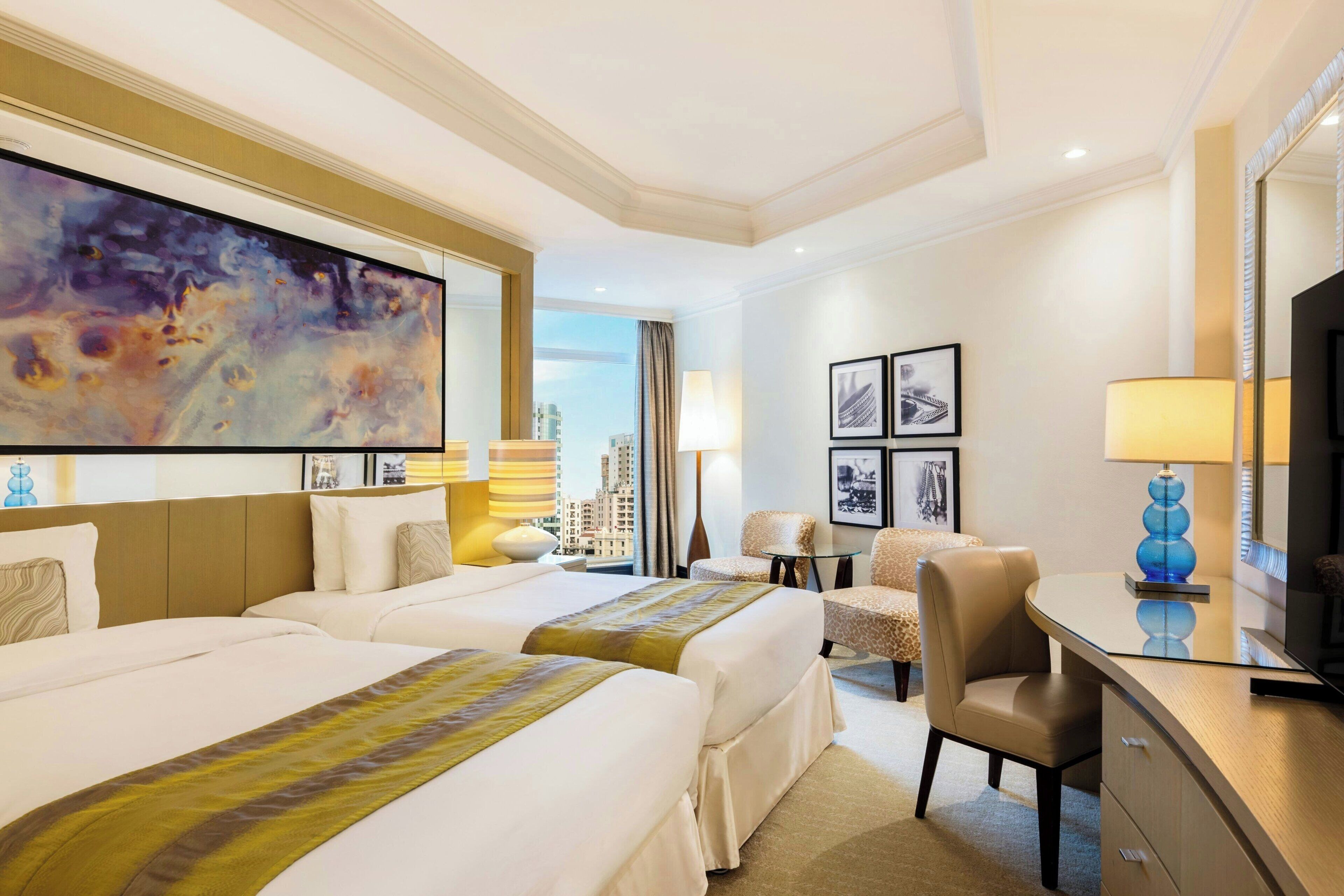 A hotel room with two beds, a desk, a chair, a TV, and a large painting on the wall.