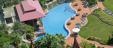 An aerial view of a resort with a large swimming pool, palm trees, and a covered area with lounge chairs.